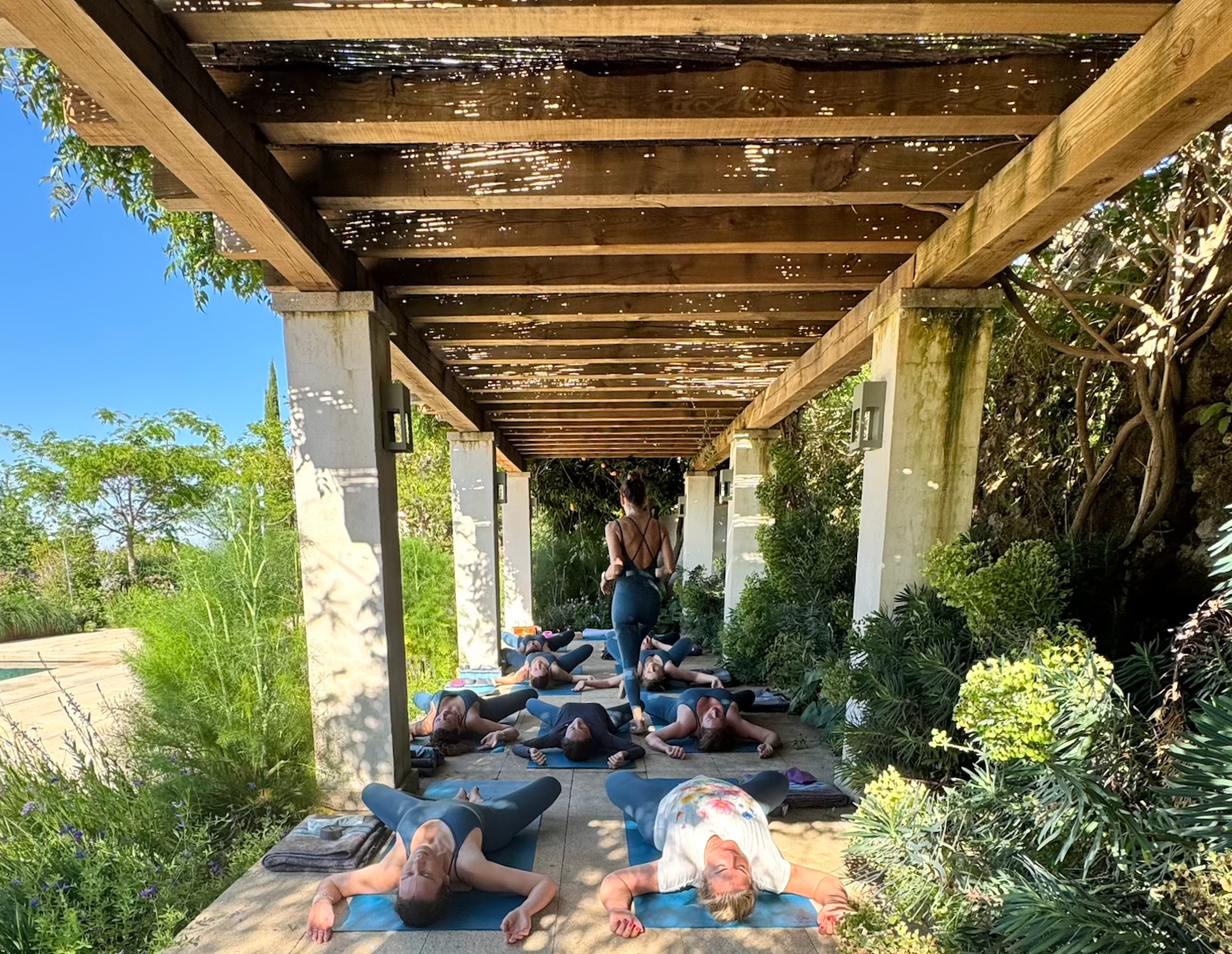 Un grupo de personas realiza una clase de yoga en un área al aire libre bajo un techo de madera, rodeado de plantas verdes y árboles.