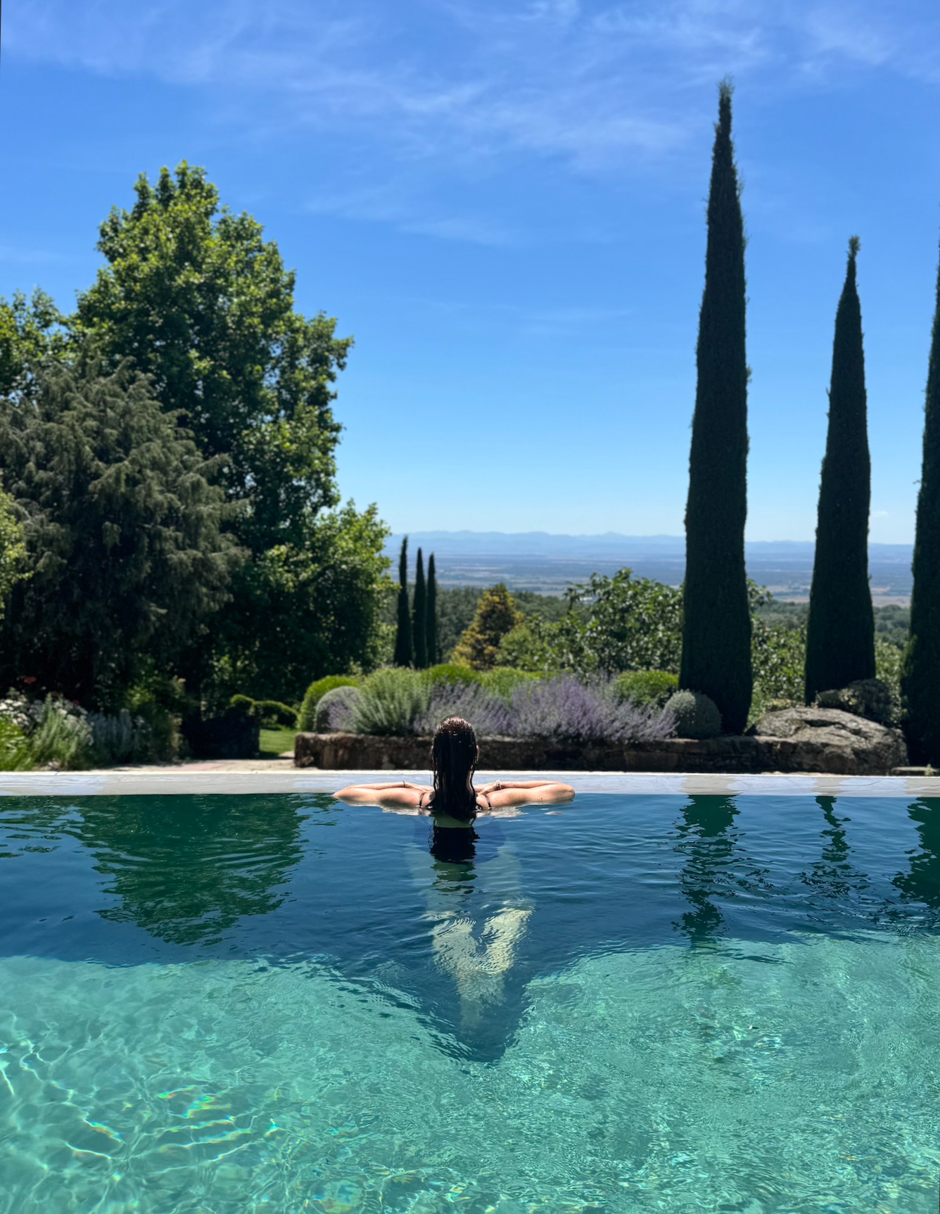 Una mujer disfrutando en una piscina con vista panorámica a un paisaje con árboles altos, césped y montañas en el fondo, en un día soleado con cielo azul.