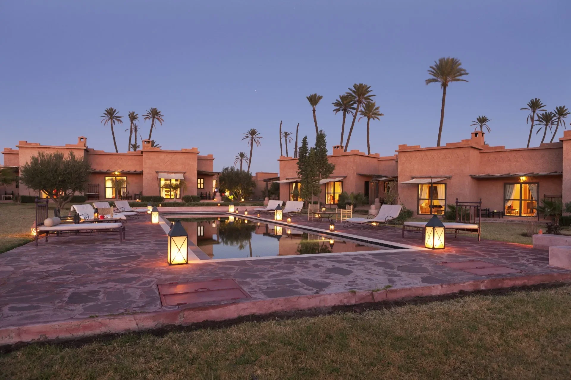 Vista de una residencia style adobe con un jardín y alberca, iluminada con lámparas y velas, rodeada de palmeras en la tarde o anochecer.