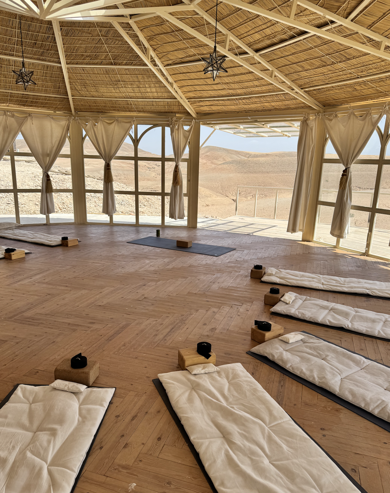 Sala de yoga con colchonetas y bloques en un espacio con ventanas grandes y vista a un paisaje desértico, con techo de caña y cortinas blancas