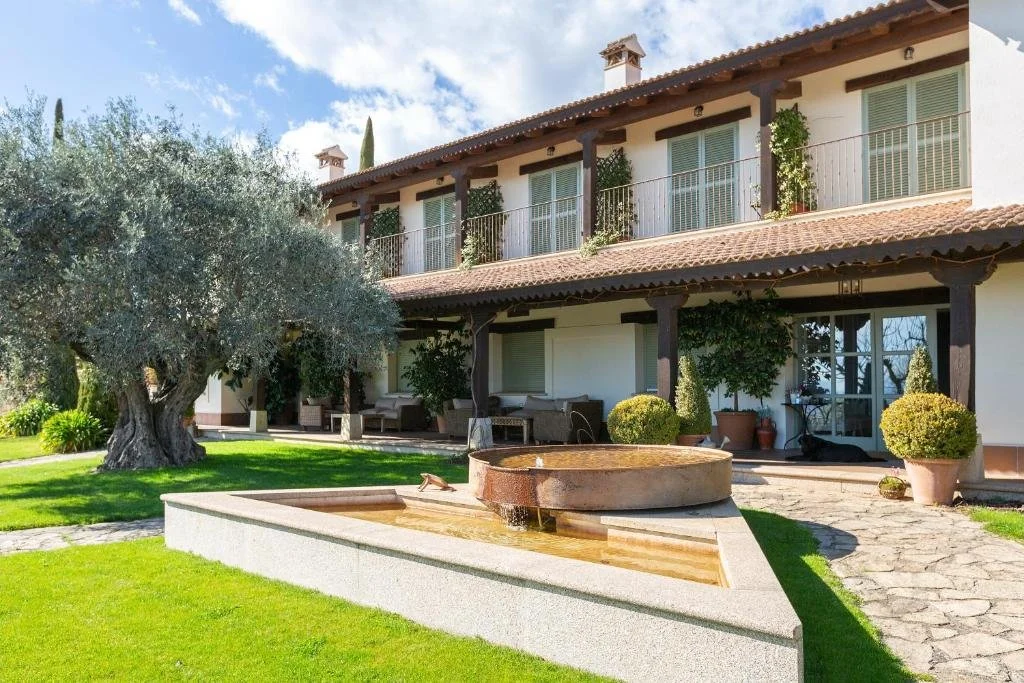 Casa tradicional de dos pisos con balcón de madera, rodeada de jardín con árboles y plantas, fuente en el centro y cielo azul con nubes.