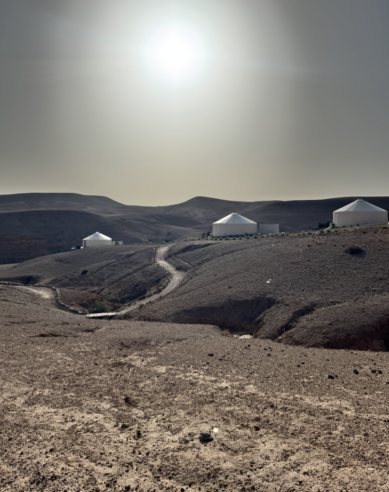 Paisaje desértico con colinas suaves y cabañas blancas de forma cónica, caminos de tierra y un sol brillante en el cielo.