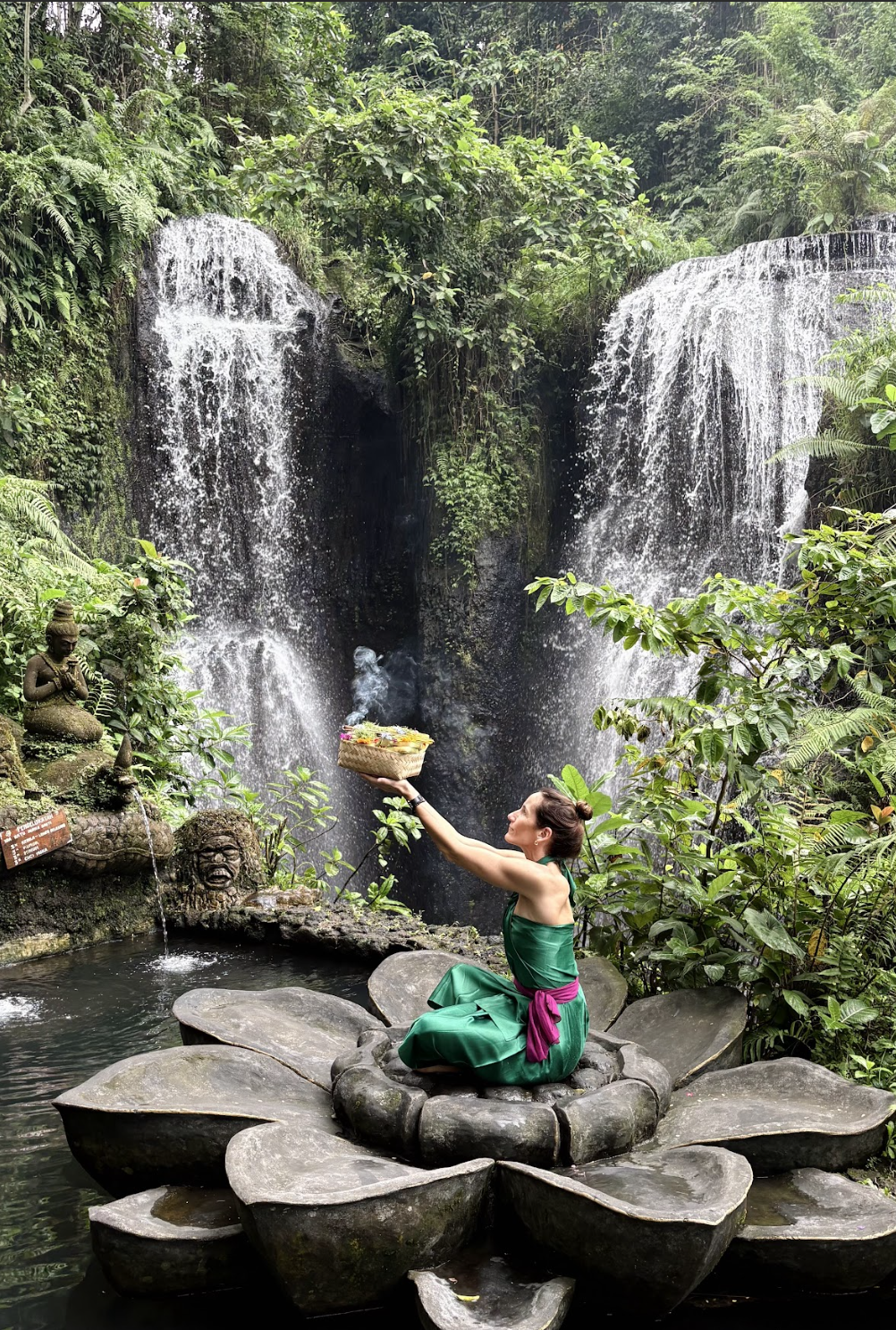 Mujer realizando una ceremonia en un altar junto a una cascada en un entorno natural tropical, rodeada de vegetación densa.