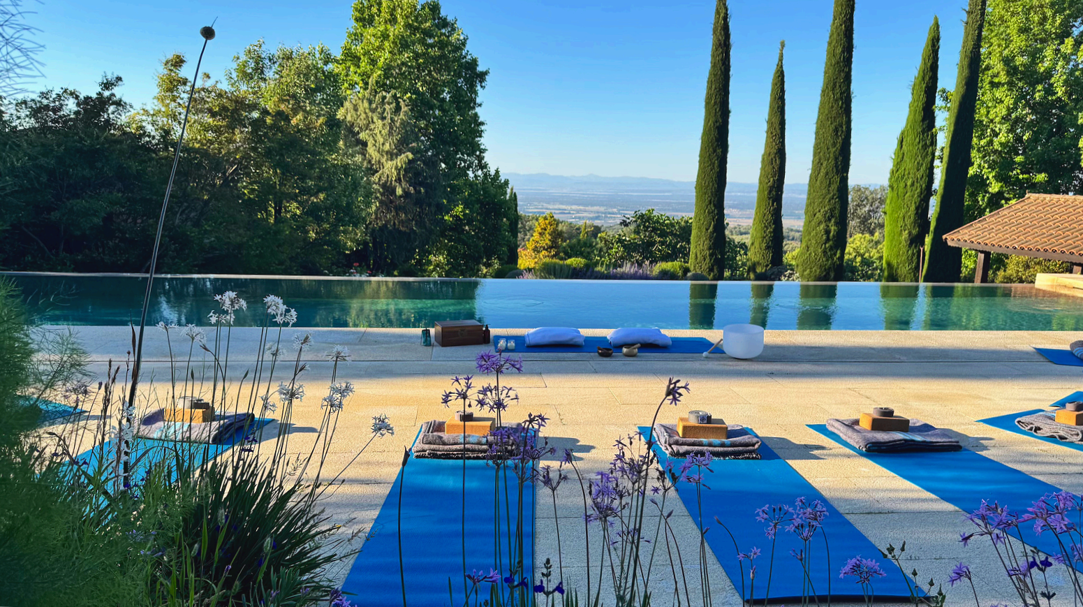 Alfombra de yoga azul y esterillas con objetos de meditación junto a una piscina, rodeada de árboles y vegetación en un entorno natural y despejado.