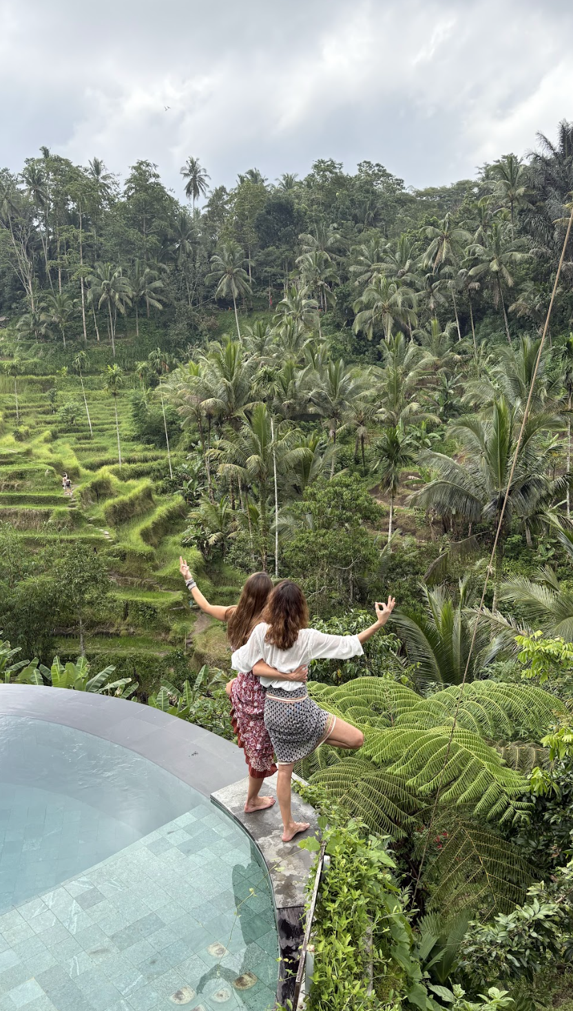 Dos mujeres jóvenes en bikini y ropa ligera posando en una terraza con vista a un paisaje de terrazas de arroz y palmeras en un entorno tropical lluvioso.