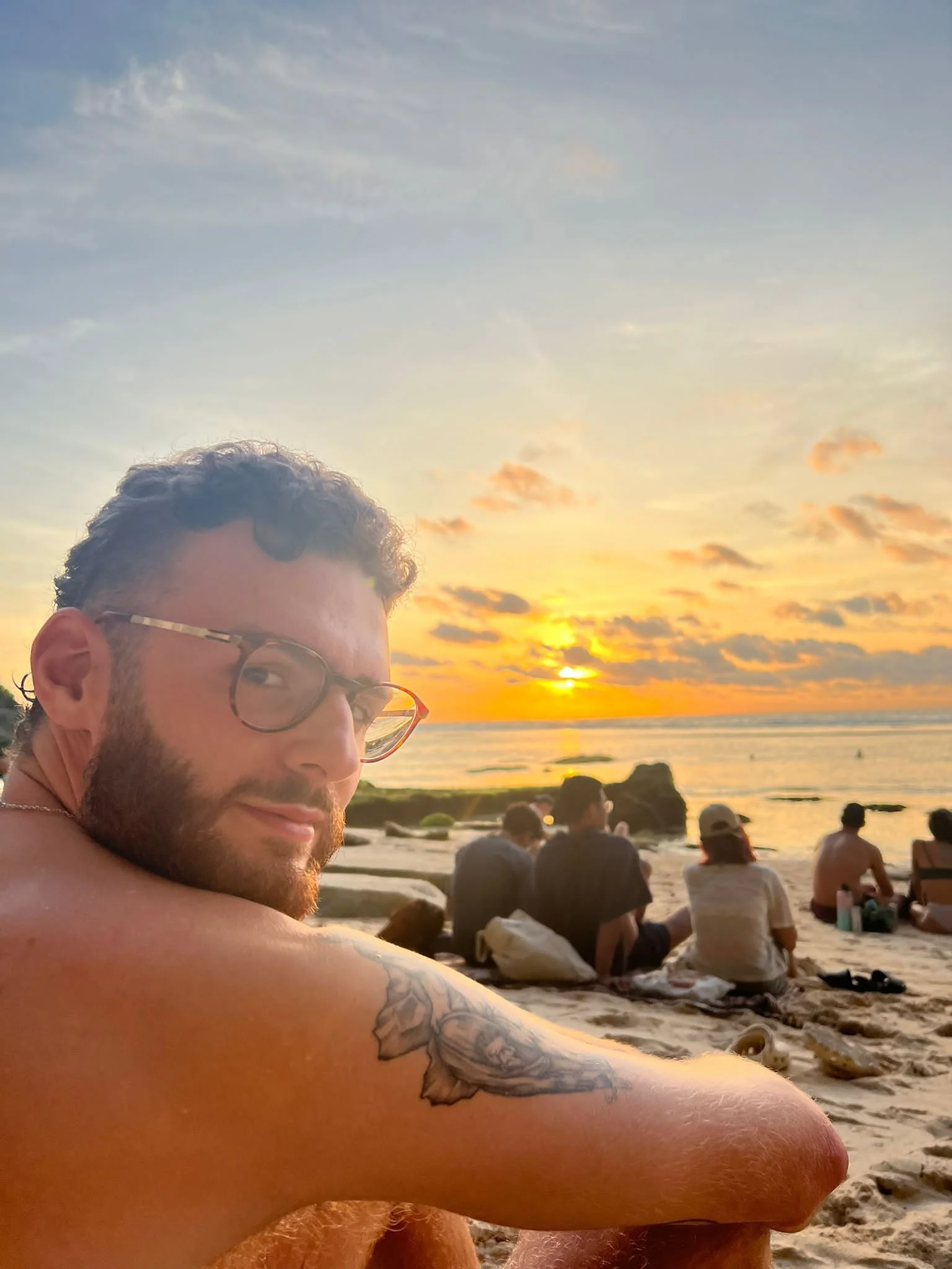 A man with glasses and a beard taking a selfie on a beach during sunset, with people sitting on the sand and the ocean in the background.