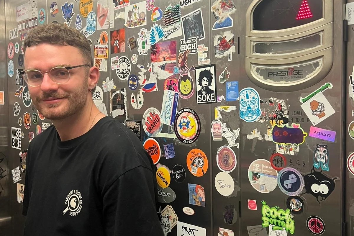 A young man with glasses, short curly hair, and a goatee, wearing a black t-shirt, standing in front of a wall covered with colorful stickers and decals.