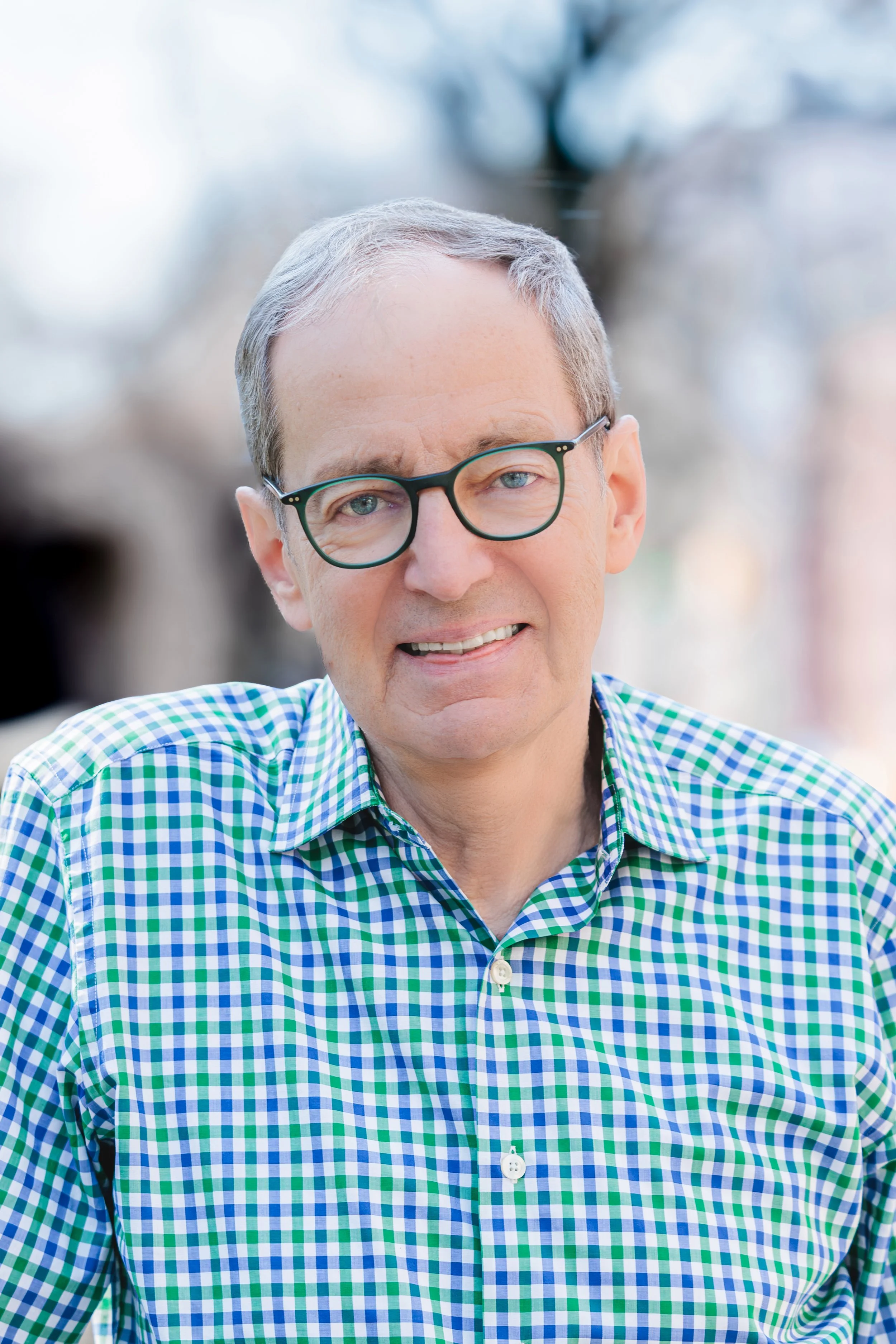 Close-up of a smiling man with glasses, wearing a blue and green checkered shirt, outdoors with blurred background.