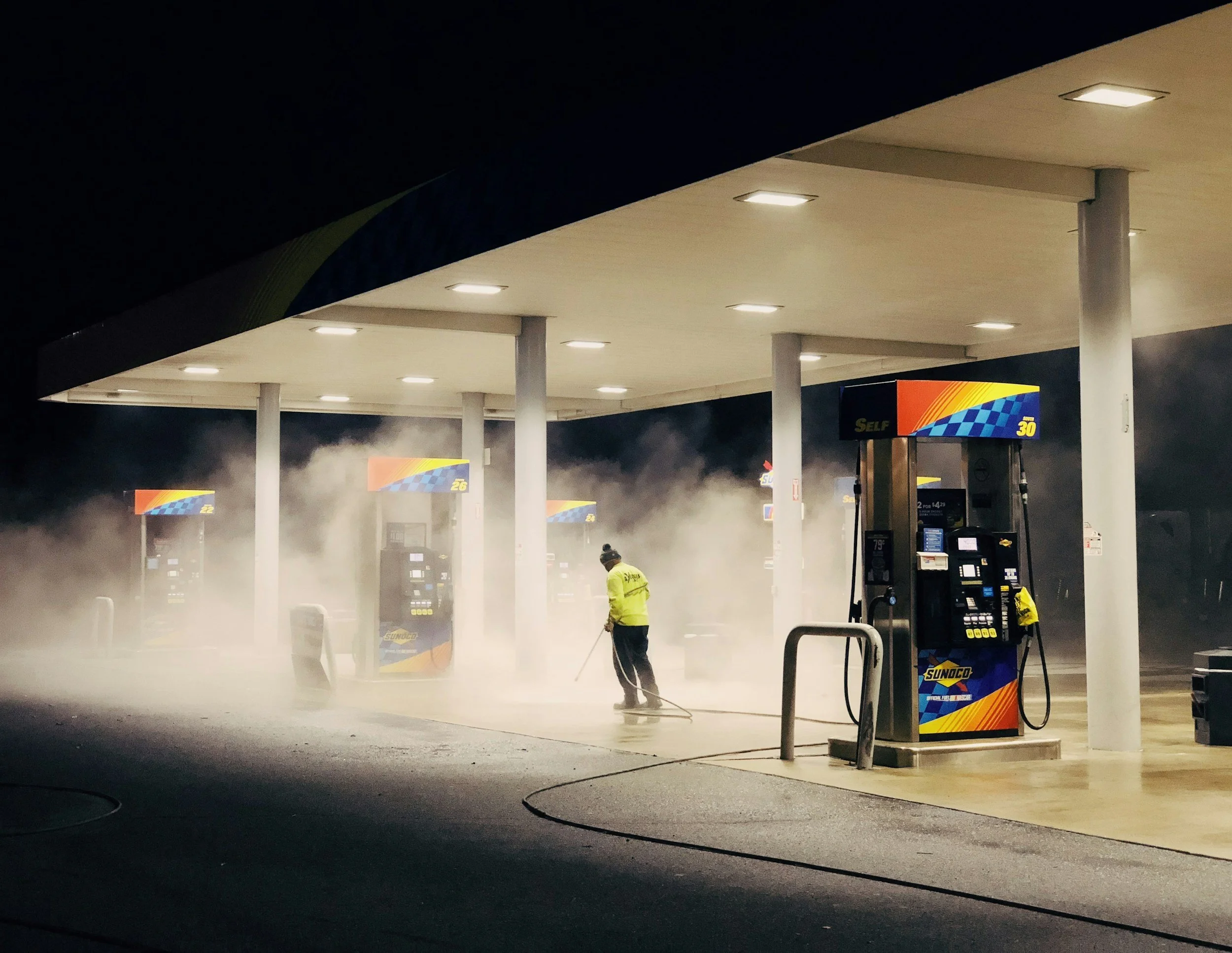 A gas station at night with steam rising from the ground. A worker in a yellow jacket is cleaning the pavement near the fuel pumps, which have bright colorful signage, including Sunoco branding.