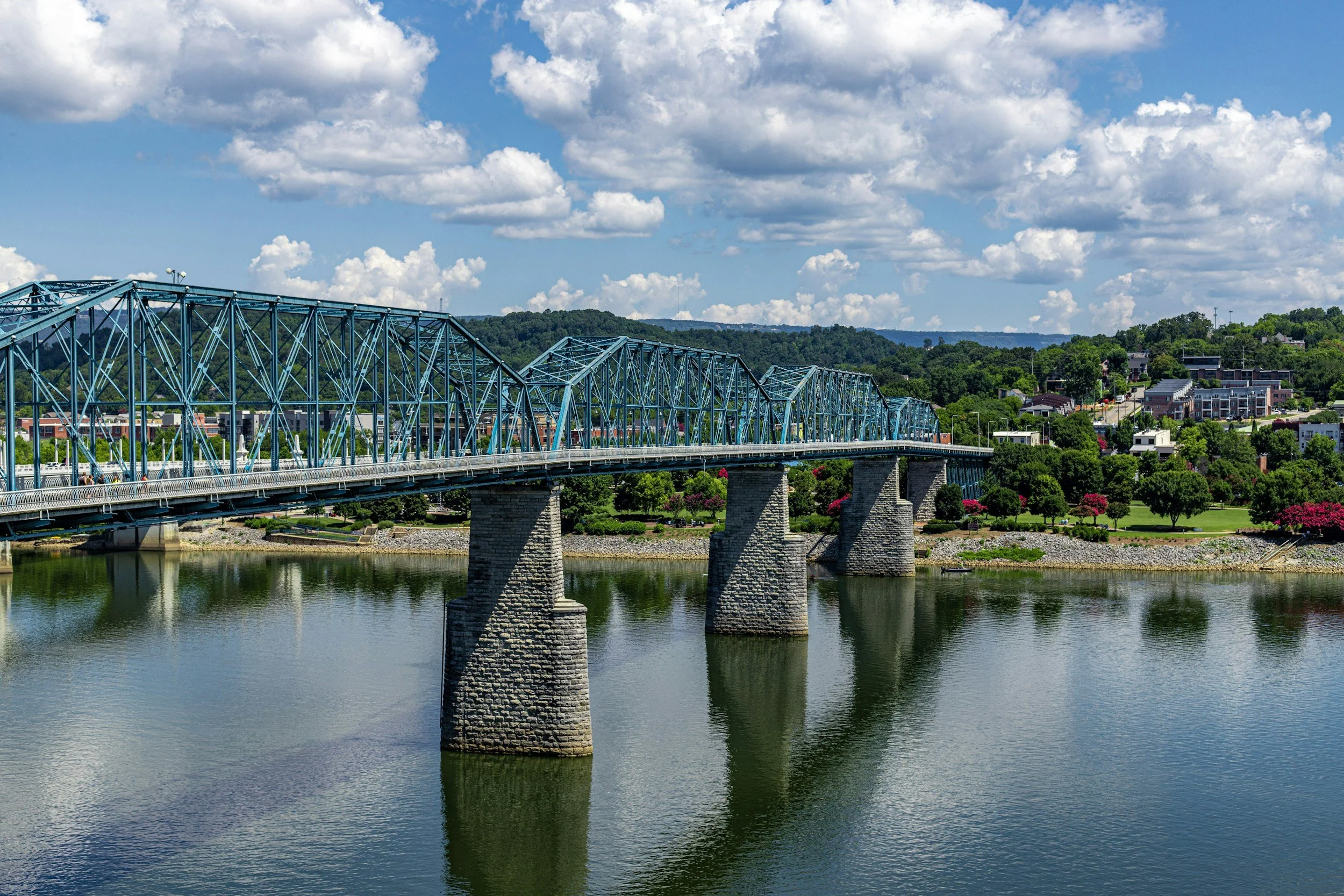 A blue steel truss bridge crossing a calm river with reflections, green hills, and a residential area with trees and colorful flowers in the background under a partly cloudy sky.