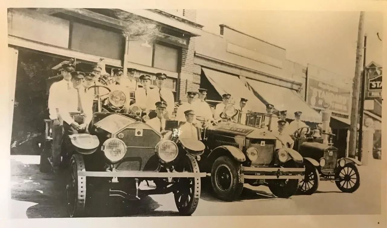 Black and white photo of a group of people standing next to vintage cars on a street in front of storefronts, with a street sign visible.