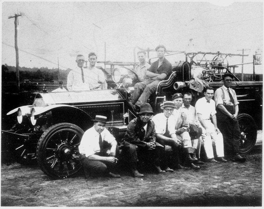 A vintage black-and-white photograph of eleven men gathered around a fire truck, some sitting on the ground and others standing, with a rural landscape in the background.