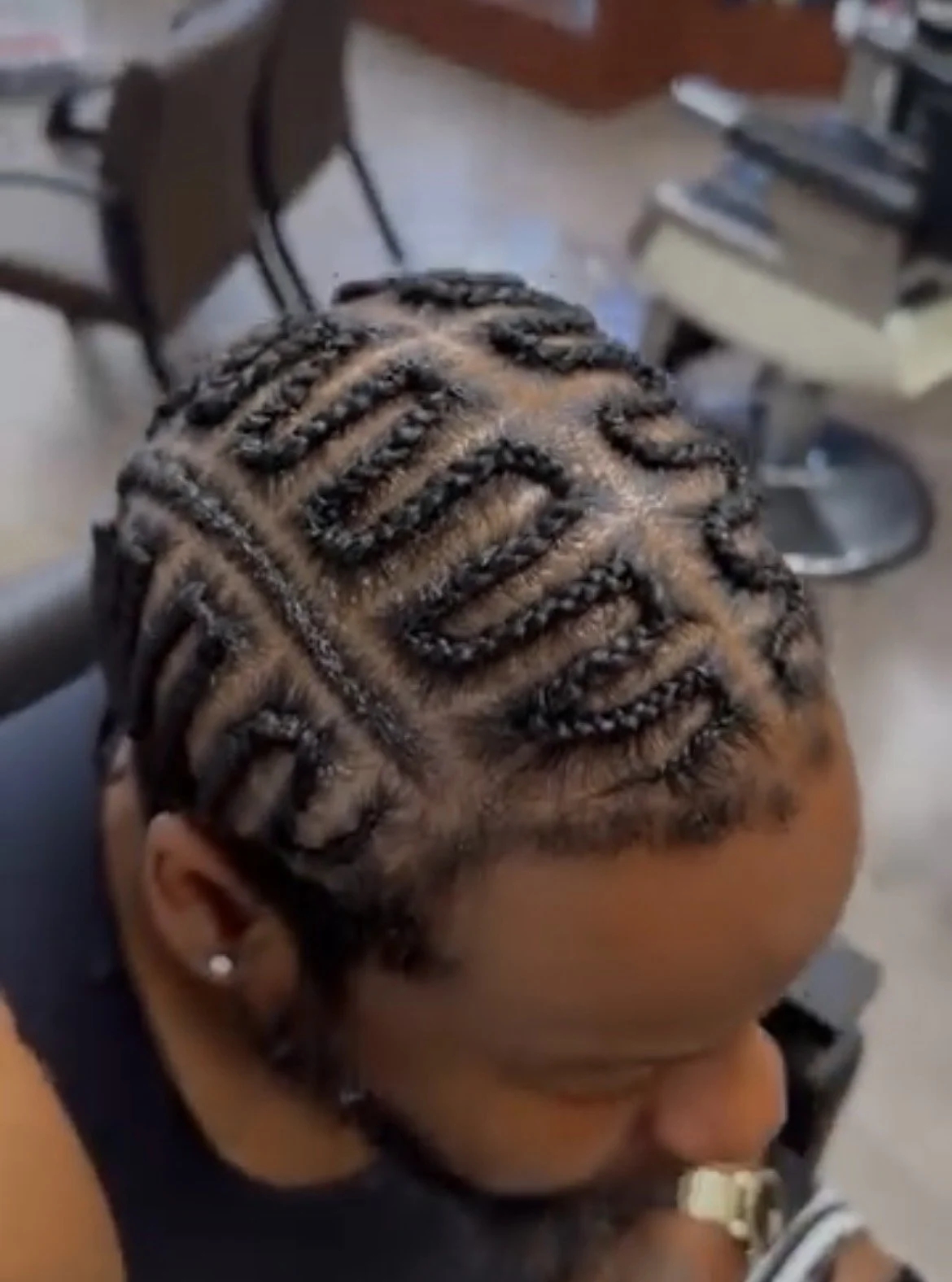 A person with freshly styled hair in tight, intricate braids with a pattern, sitting in a hair salon with salon chairs and shelves with books in the background.