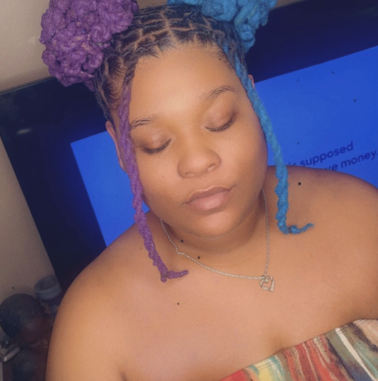 A young woman with purple and blue braided hair, eyes closed, wearing a necklace with a heart pendant, and sitting in front of a television screen.