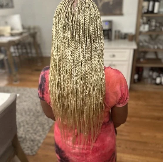Back view of a woman with long blonde box braids, wearing a tie-dye pink and black shirt, in a cozy home kitchen.