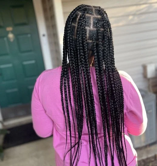 Back view of a woman with long braided hairstyle wearing a pink shirt, standing outside a house with a green door.