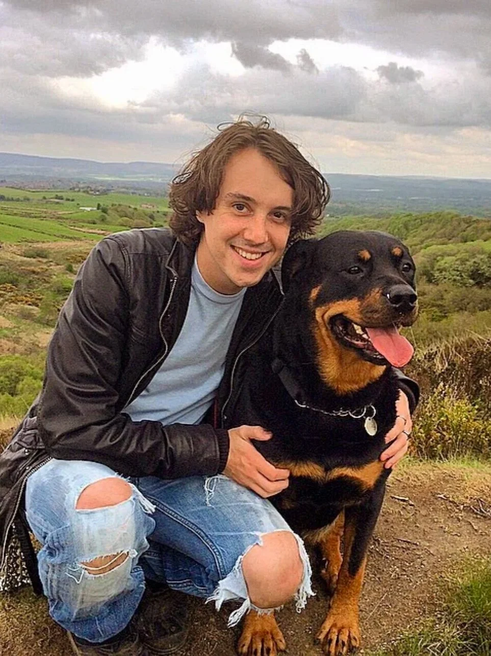 A young man with curly brown hair smiling and hugging a large Rottweiler dog outdoors on a hilltop, with green fields and cloudy sky in the background.