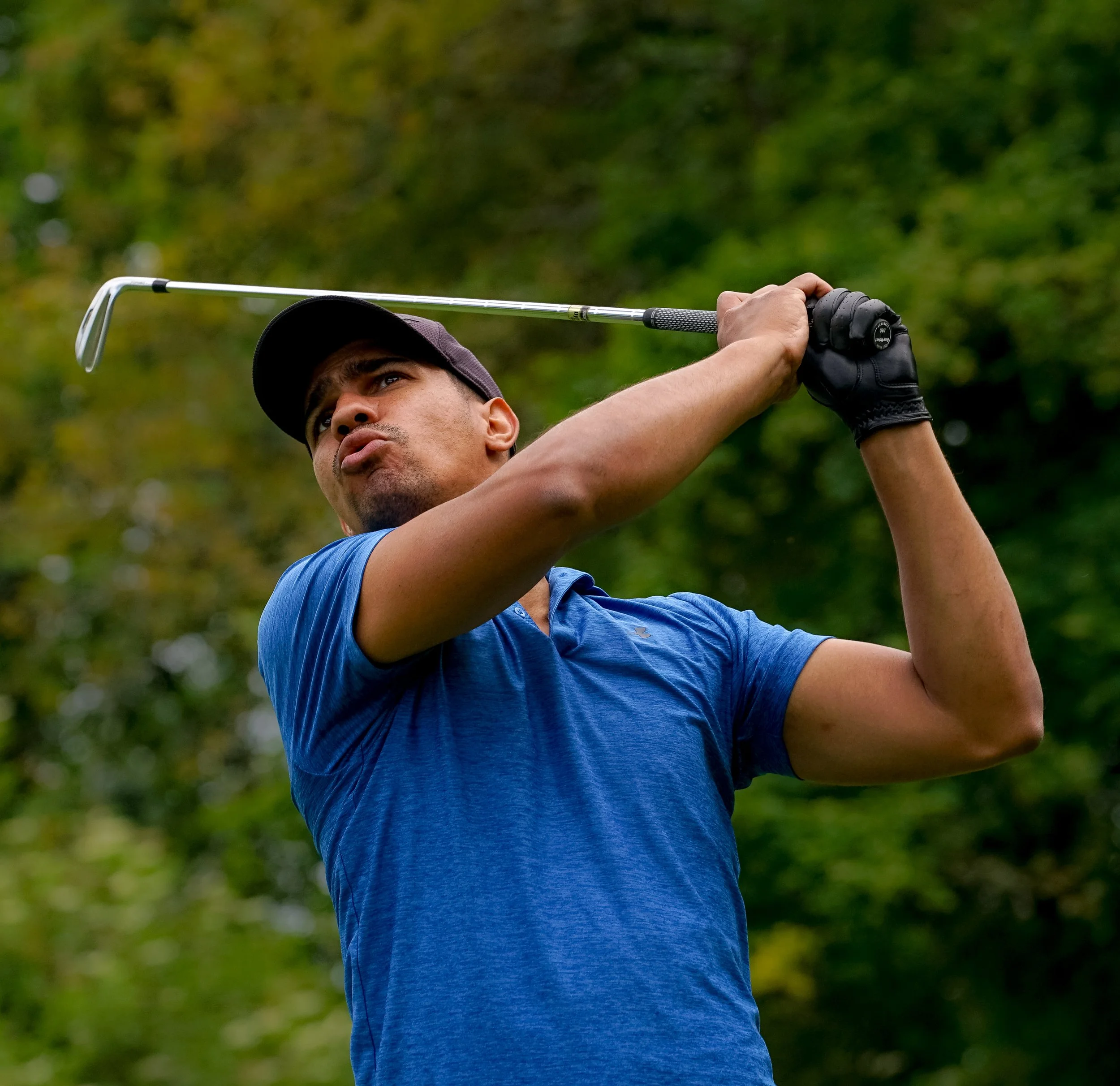 Un homme en t-shirt bleu, portant une casquette noire, joue au golf en levant un club de golf en direction de la balle, avec un fond de feuillage vert
