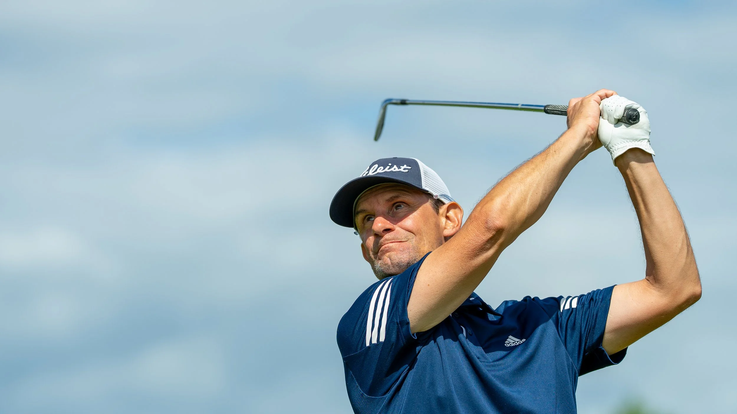 Un golfeur en pleine frappe avec un club de golf, portant une casquette et un gant blanc, en extérieur sous un ciel clair.