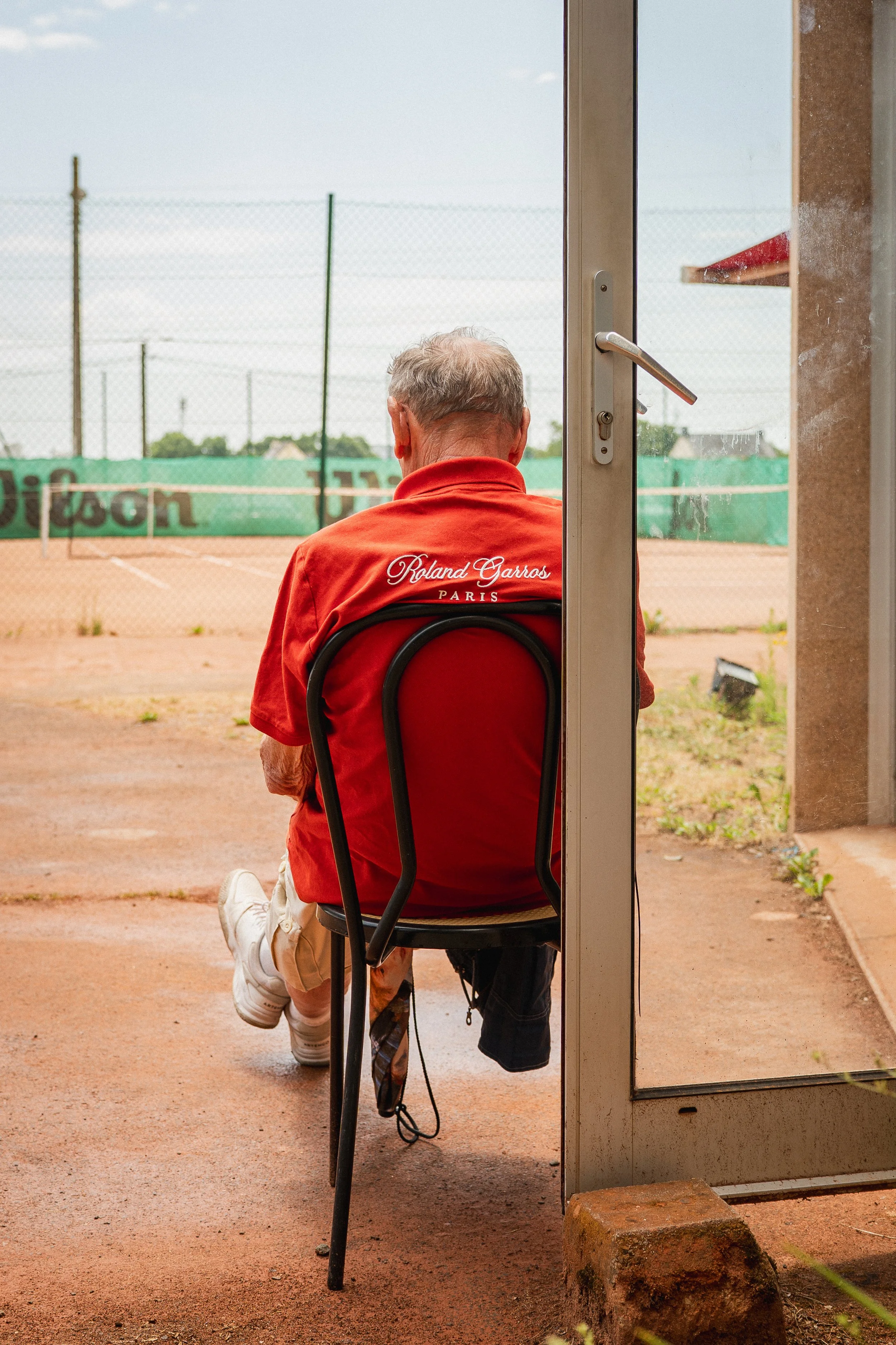 Un homme assis sur une chaise, vu de dos, regarde un terrain de tennis. Il porte un t-shirt rouge avec l'inscription 'Roland Garros PARIS' au dos.