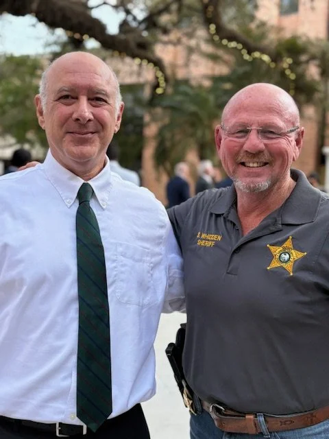 Two men stand side by side outdoors, smiling. The man on the left wears a white shirt and green striped tie, while the man on the right is in a Sheriff's uniform with a badge and glasses. Background includes trees and string lights, indicating an outdoor event.