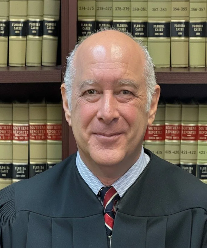A judge Robert Ostrov wearing a black robe and a striped tie, standing in front of shelves filled with legal books in a law library.