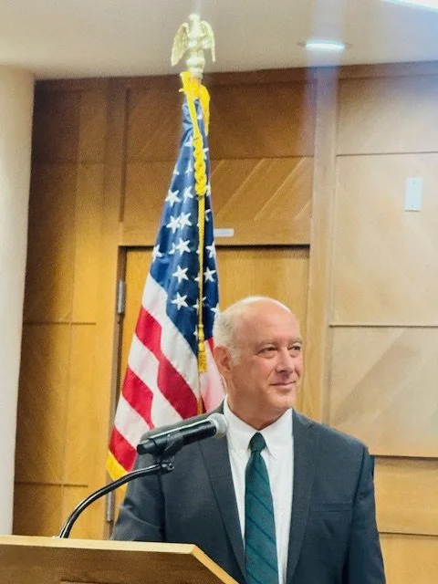 Robert Ostrov in a suit and tie standing at a podium with a microphone, behind him a wooden wall, and an American flag with a golden eagle finial on top.