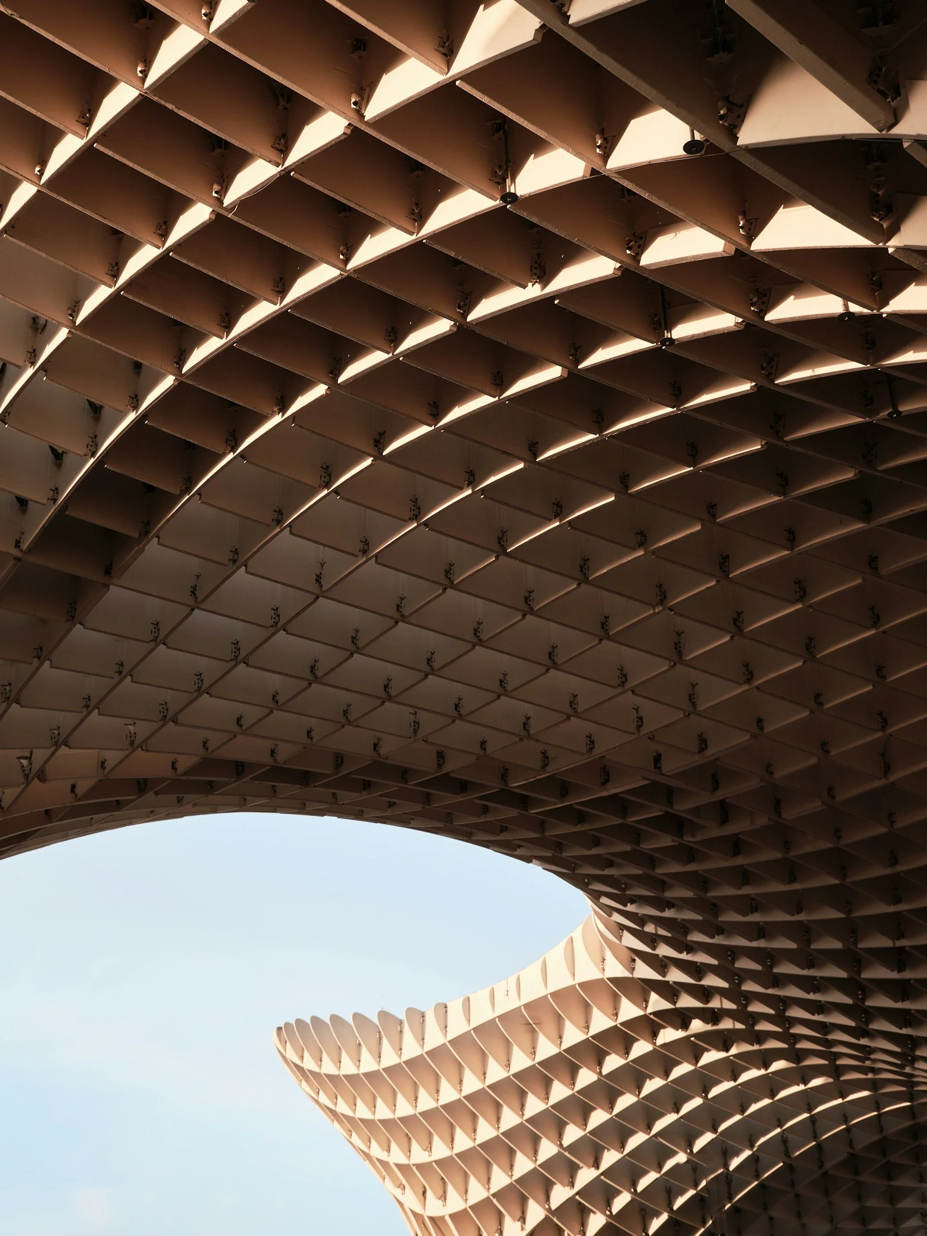 Close-up view of an architectural structure with a parametric, curved design made of beige-colored panels, creating a pattern against a clear blue sky.