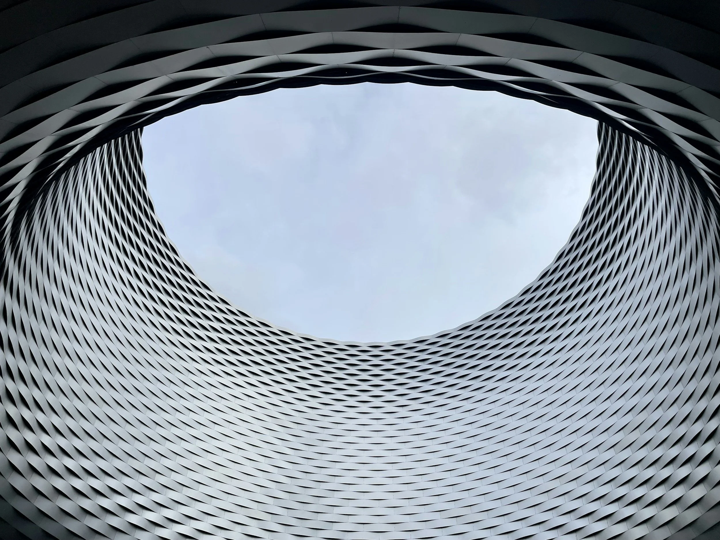 Looking up at a modern building with a circular, twisting design and an opening in the center, with a cloudy sky in the background.
