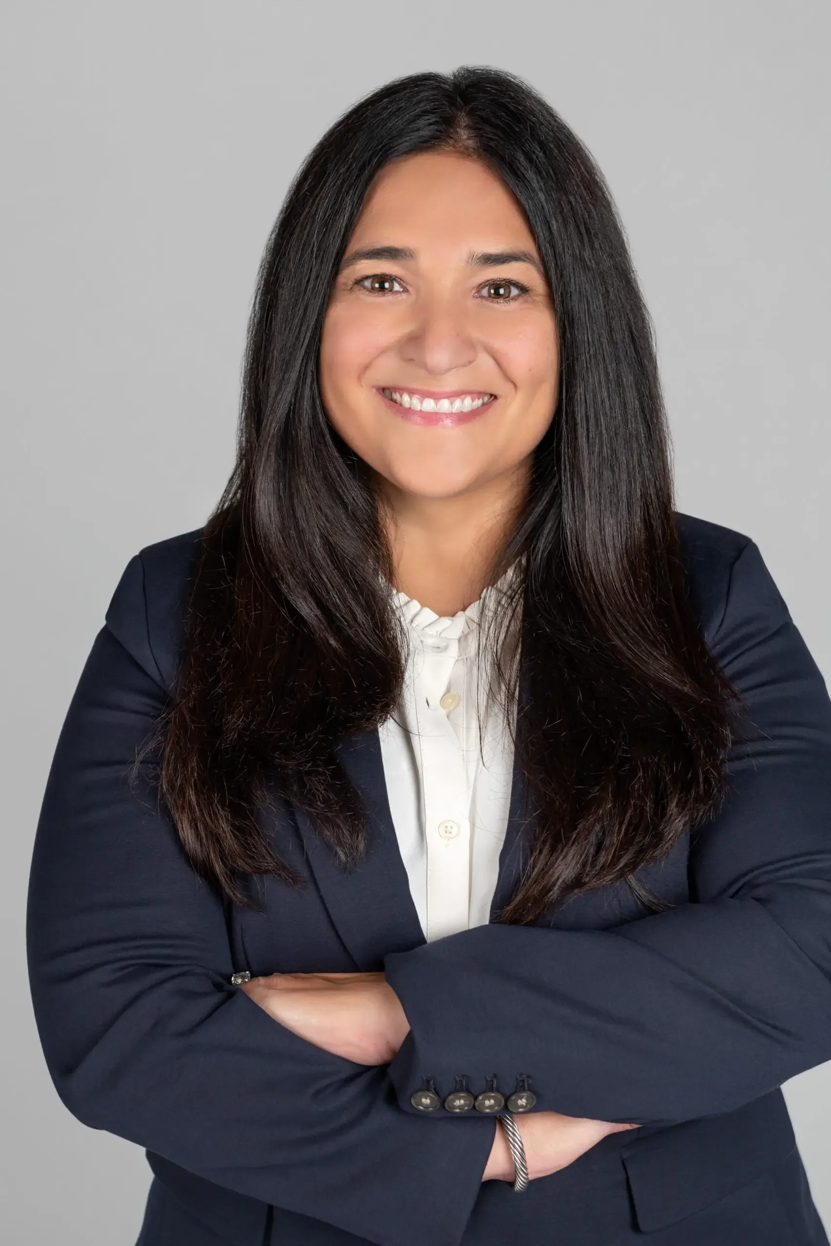 A professional woman with long dark hair, wearing a dark blazer, white blouse, smiling with arms crossed against a gray background.