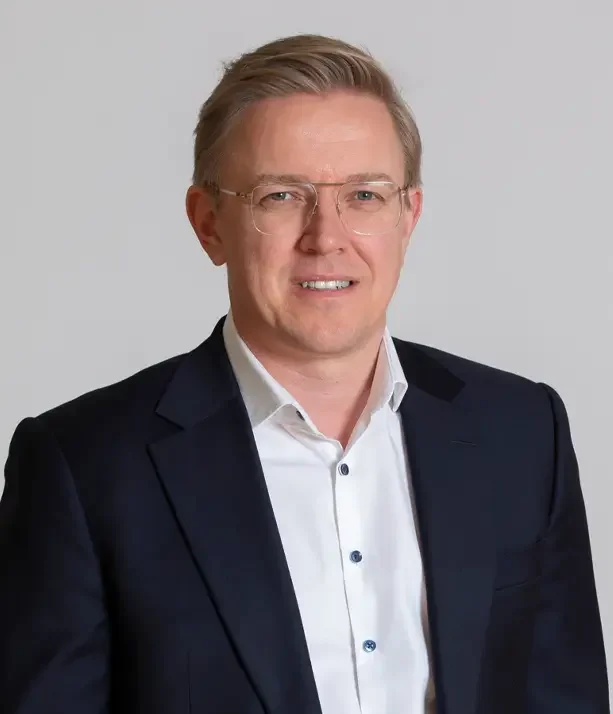 A professional man with blonde hair and glasses wearing a white shirt and dark blazer, smiling at the camera against a plain background.