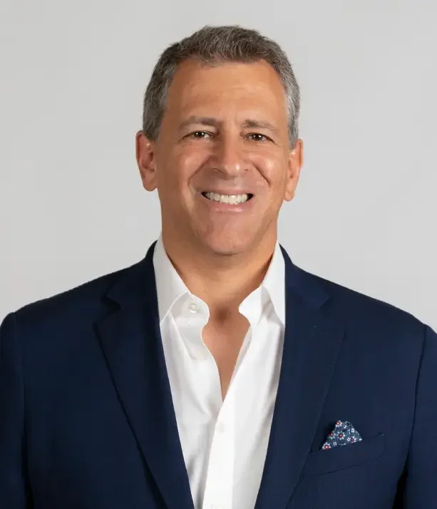 A smiling middle-aged man wearing a navy blazer, white dress shirt, and a pocket square, standing against a light gray background.
