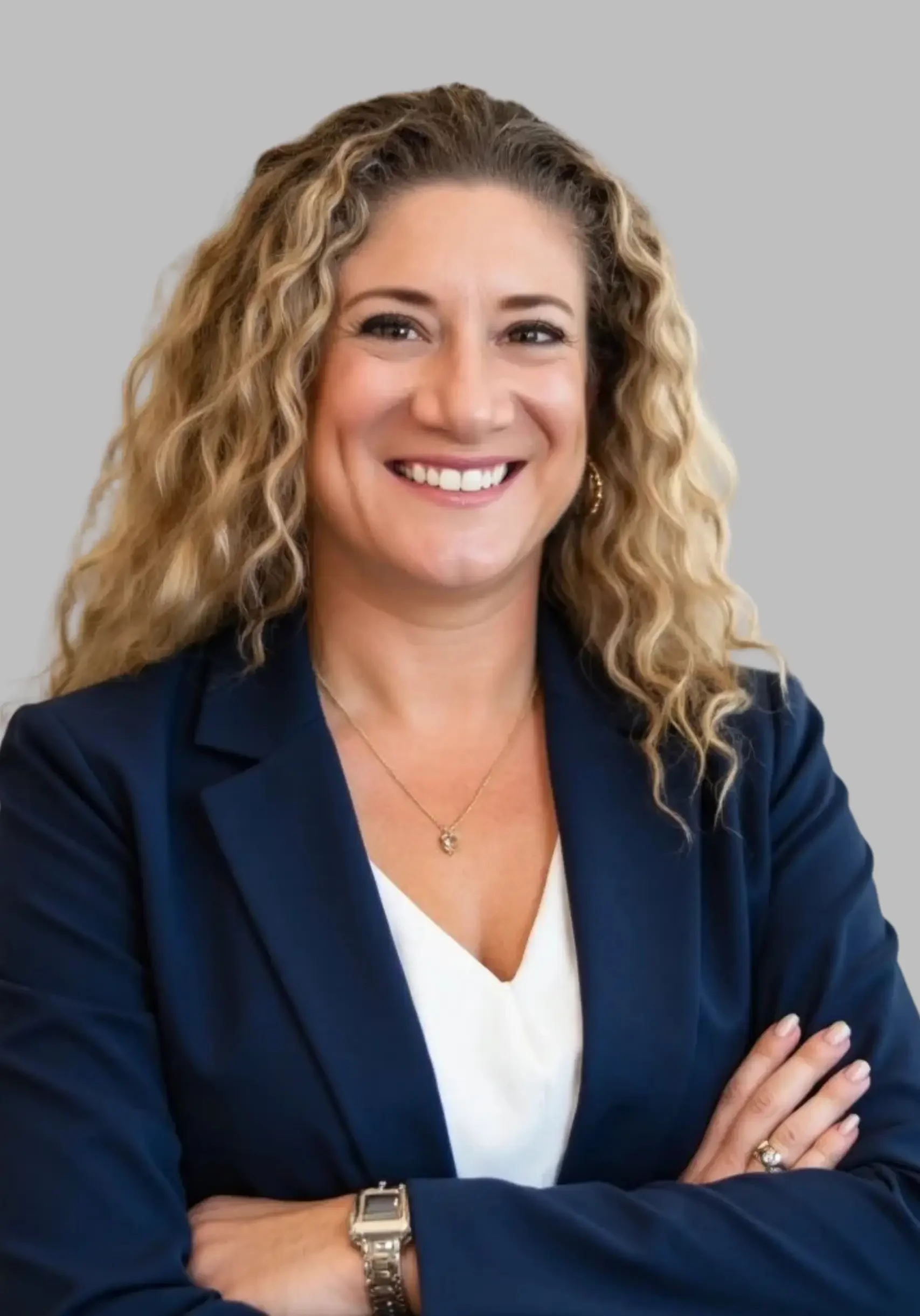 A woman with curly blonde hair smiling, wearing a dark blazer and a white top, with her arms crossed, set against a light gray background.