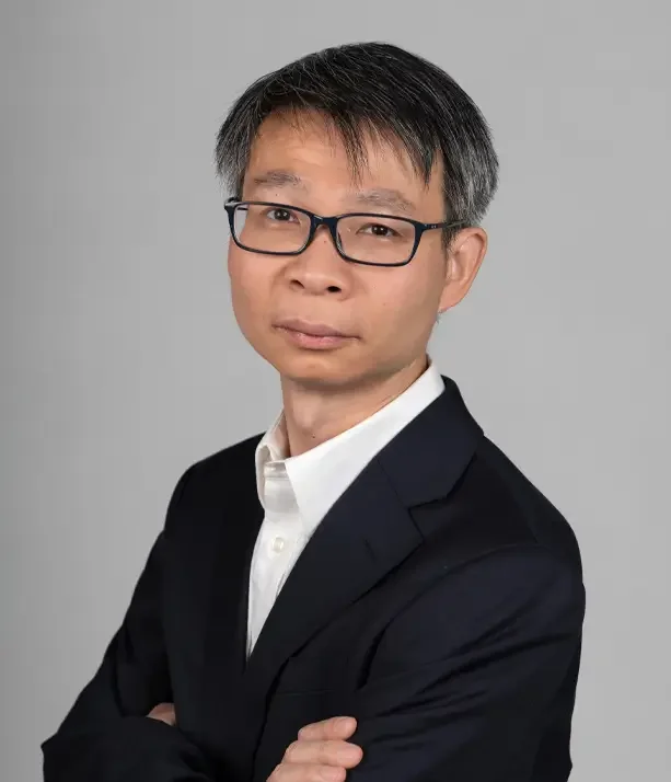 Portrait of a man with glasses, wearing a dark suit and white shirt, standing against a gray background.