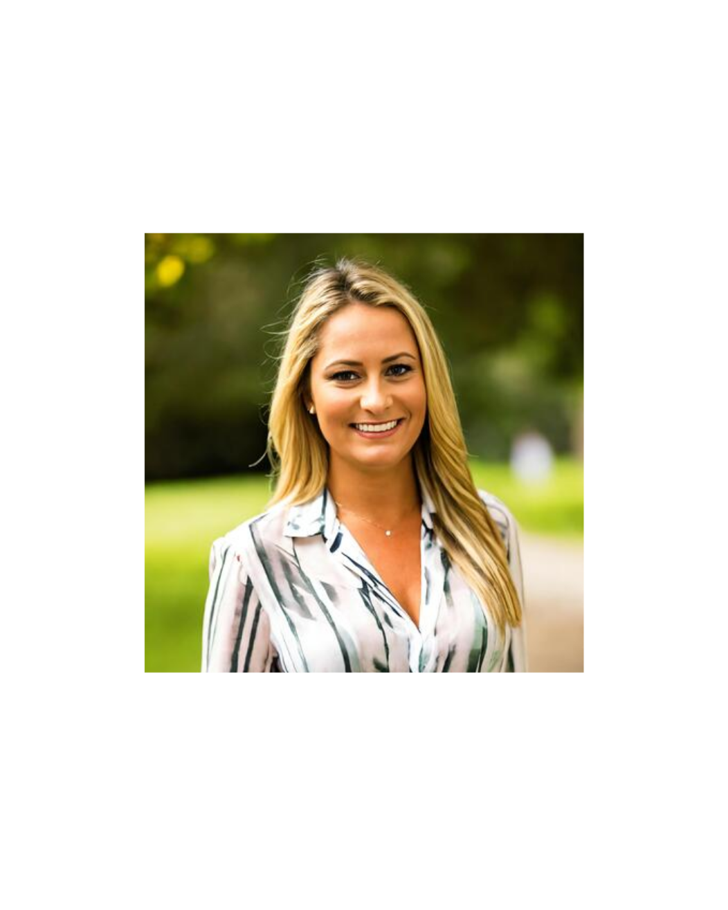 A smiling woman with long blonde hair, wearing a white blouse with black and gray stripes, standing outdoors in a park with green grass and trees in the background.