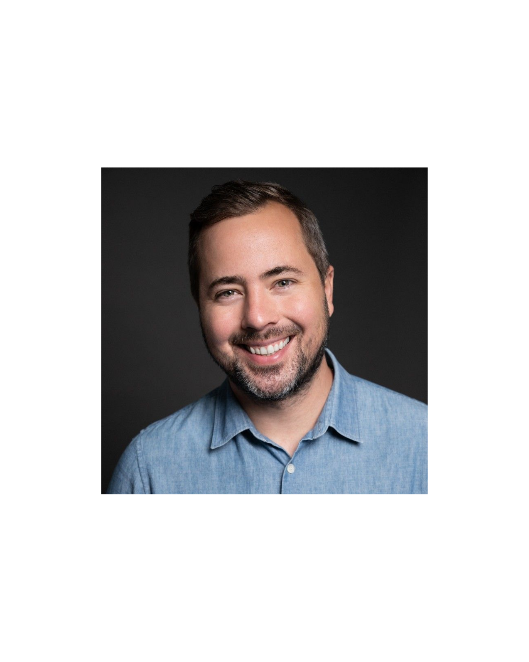 A smiling man with short brown hair and a beard, wearing a light blue button-up shirt, against a dark gray background.