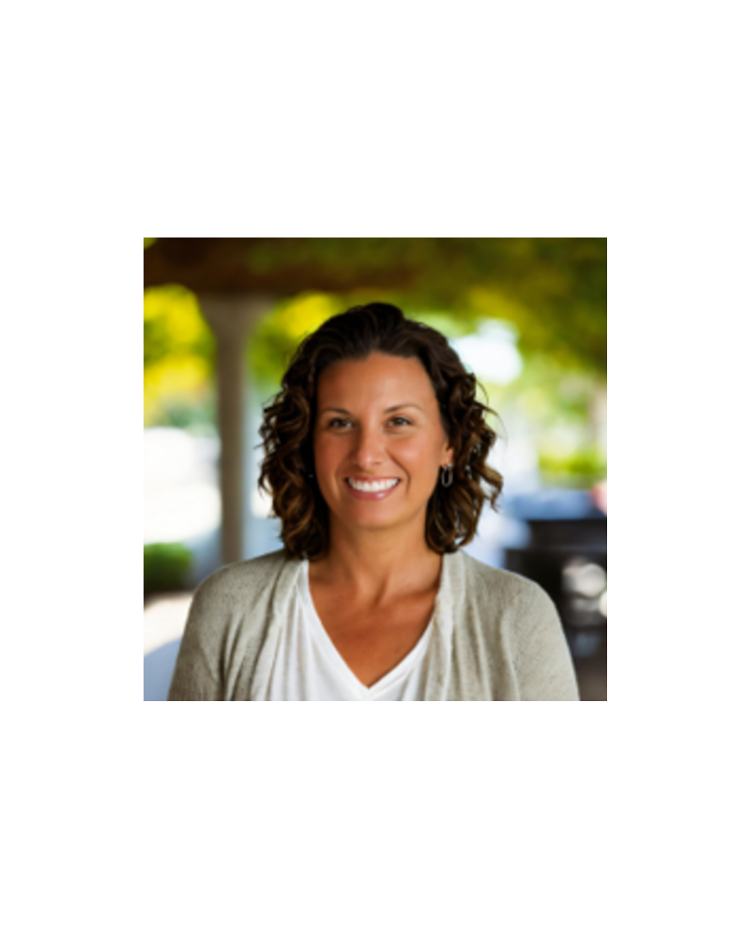 Smiling woman with curly brown hair, wearing a white top and light cardigan outdoors with blurred green trees in the background.