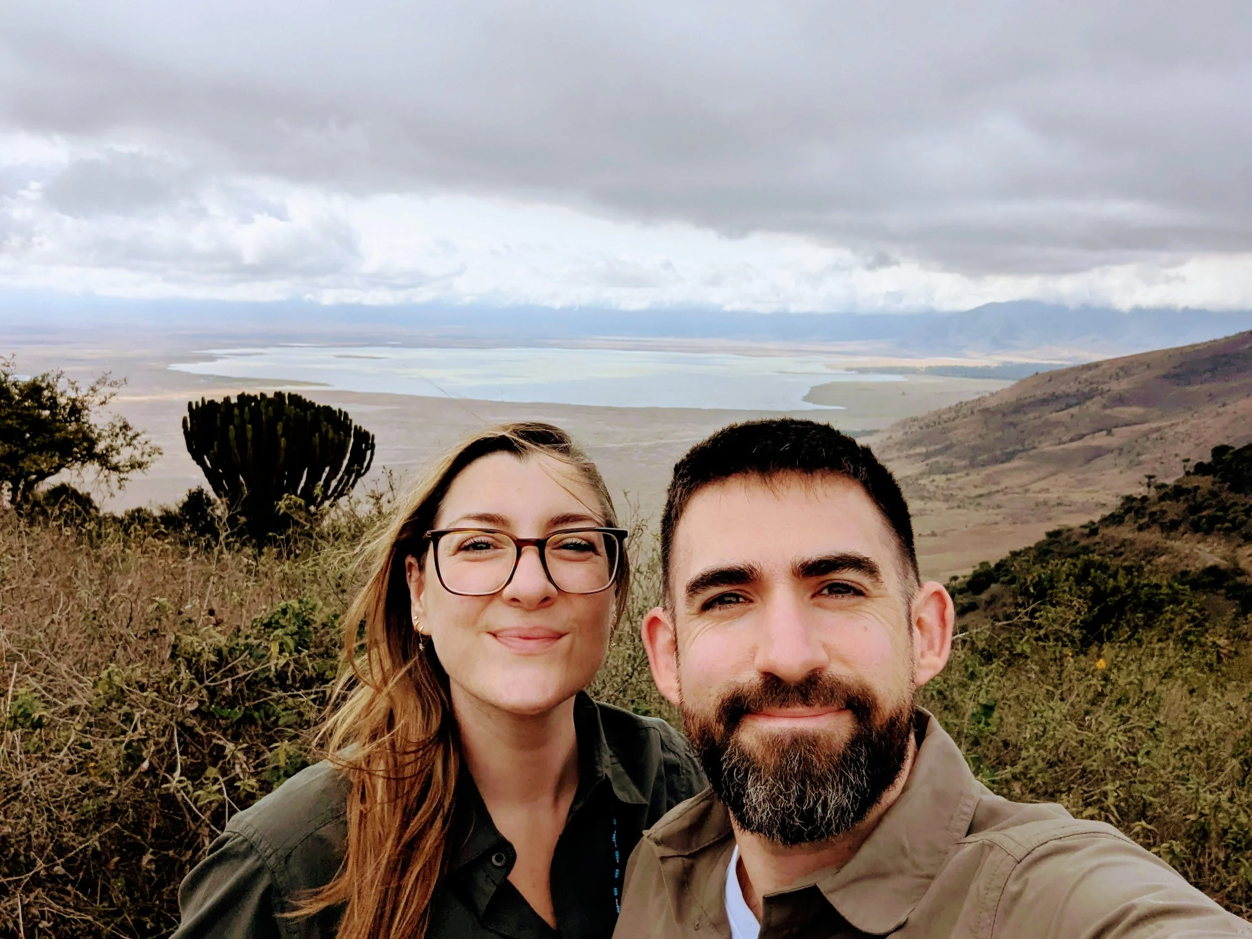 Close-up of a smiling couple outdoors with a landscape of rolling hills, water, and overcast sky in the background.