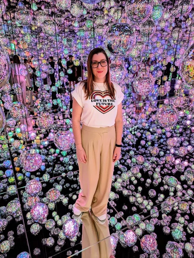 Jenny, the founder of Cantrell Consulting, standing in an immersive art installation with hanging mirrored spheres reflecting rainbow colors.