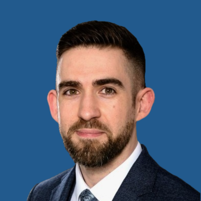 Professional headshot of a man with short dark hair, a beard, wearing a suit and tie, against a blue background.