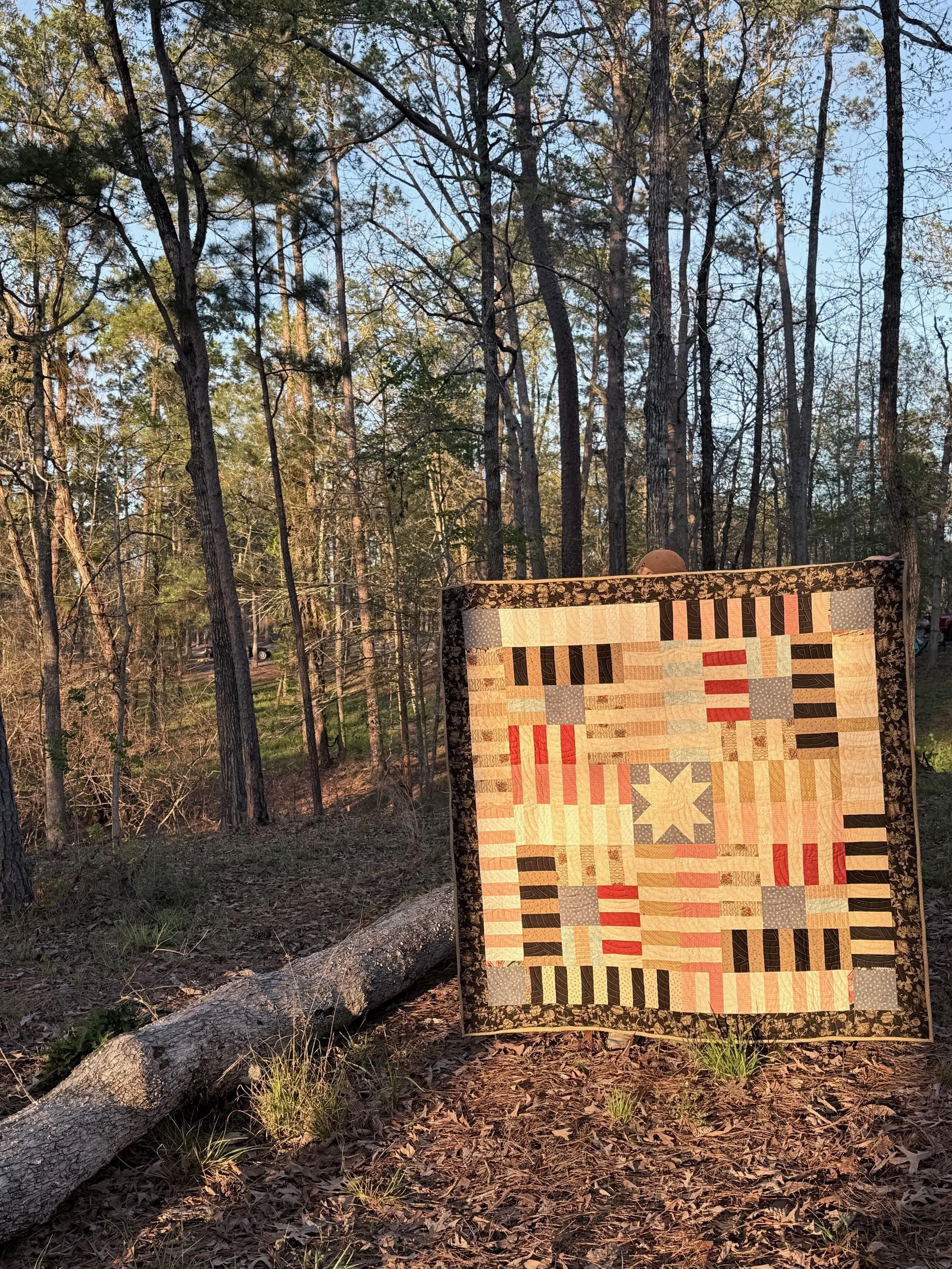 A quilt with a patchwork design featuring various geometric patterns and colors, hanging outdoors in a wooded area during daylight.