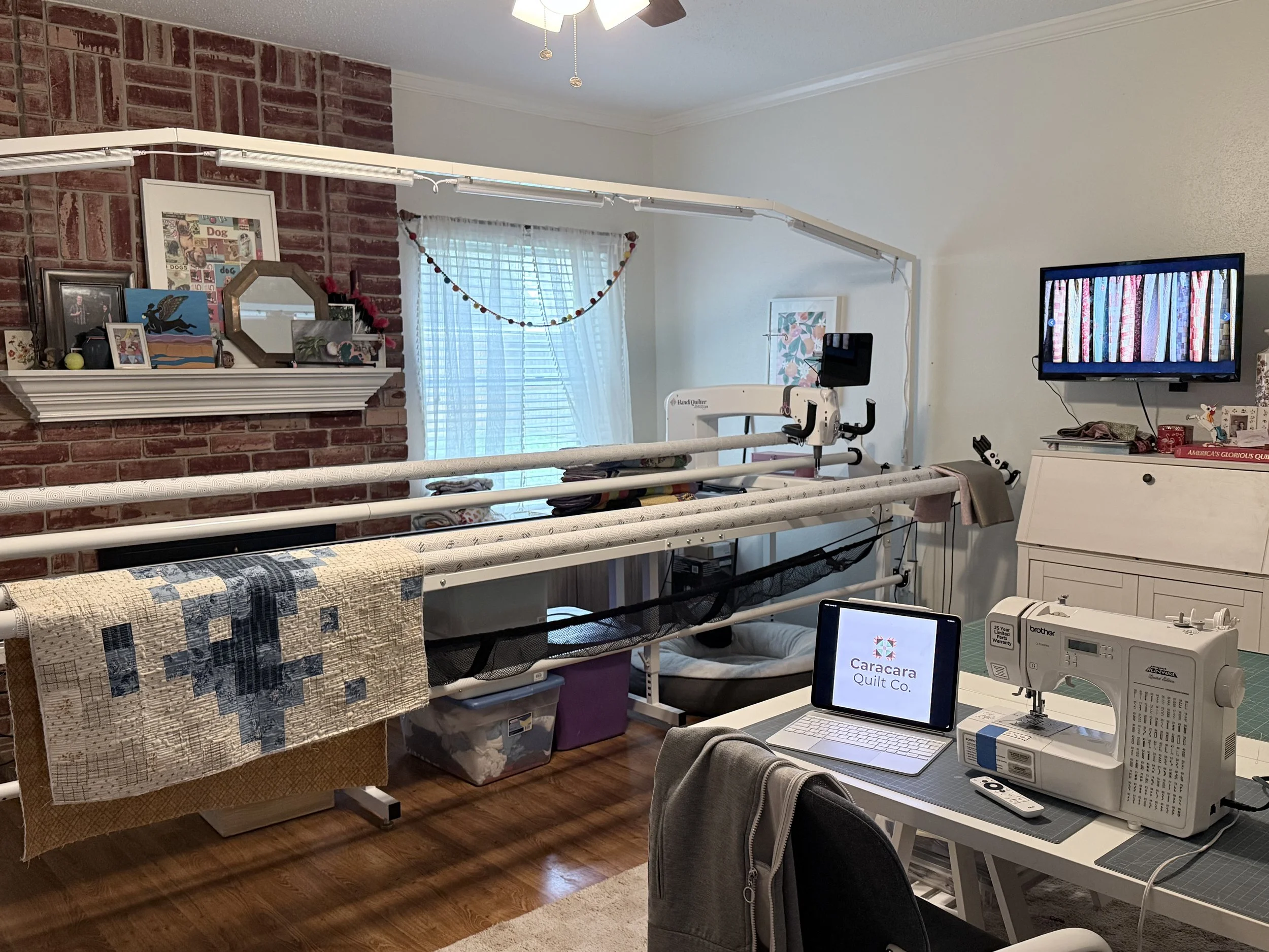 A cozy craft room with a brick wall and white walls. A window with sheer white curtains has a colorful garland hanging above it. The room contains a white sewing or quilting machine on a desk, a monitor displaying a logo for Caracara Quilt Co., a white cabinet, and various artwork and decorative items on the fireplace mantle and walls. A quilt in progress hangs over a table, and sewing supplies and a computer are on the desk.