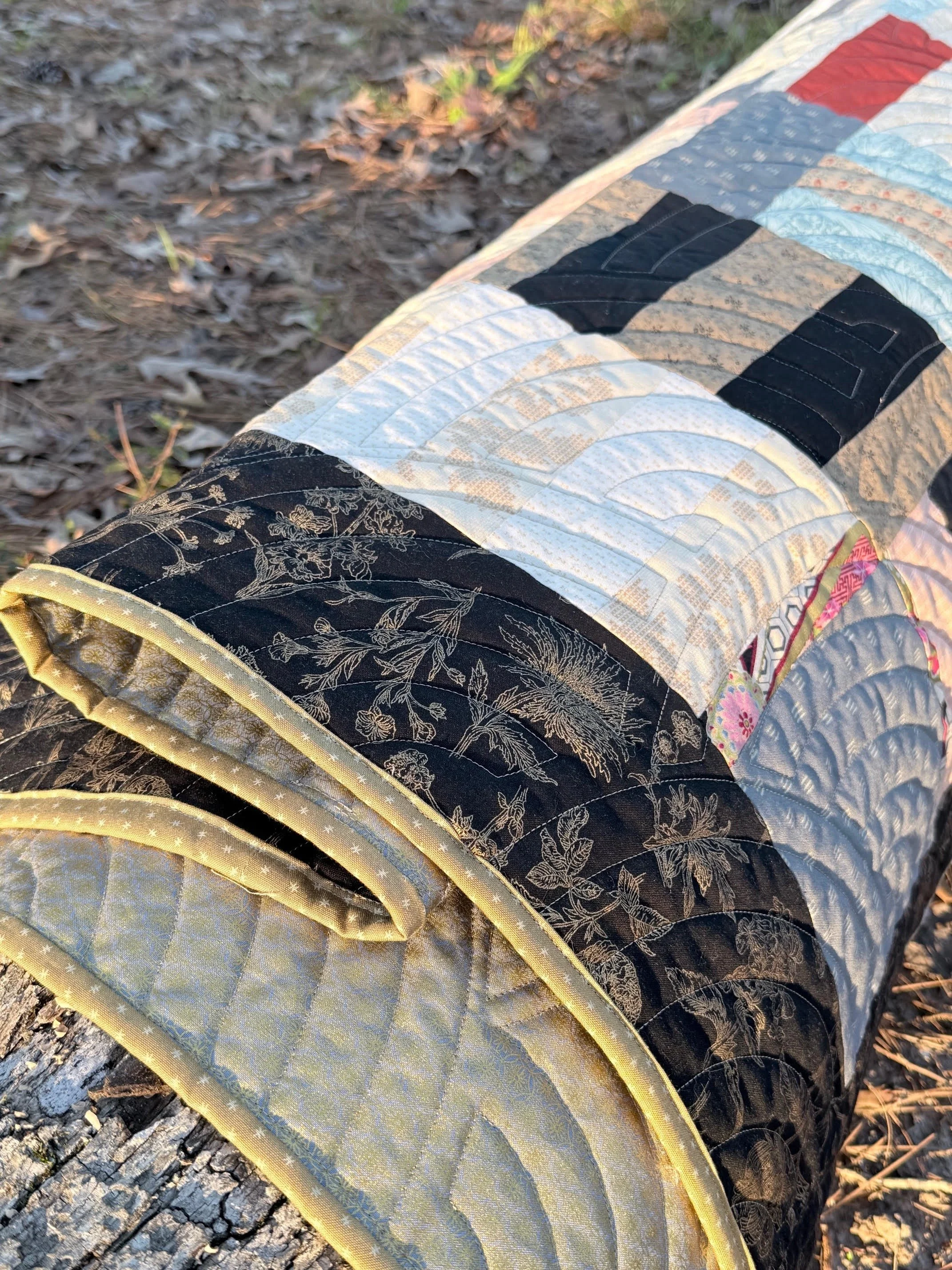 Colorful quilt with various patterns and colors, draped over a surface outdoors near dried leaves and grass.