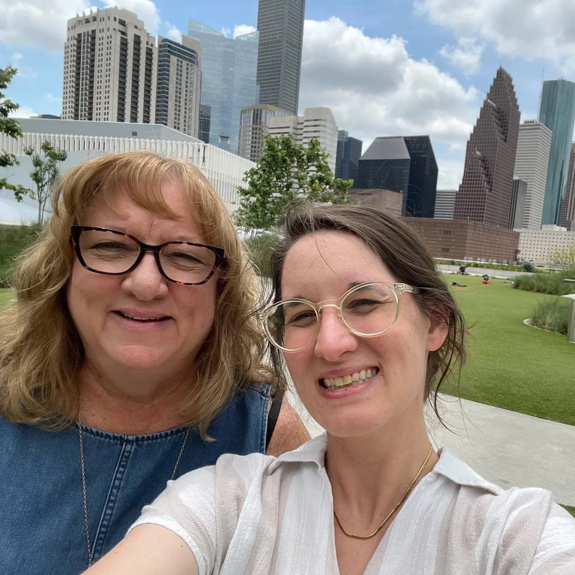 Two women smiling outdoors in front of a city skyline with tall buildings and partly cloudy sky. One woman has blonde hair and wears glasses, the other has brown hair and also wears glasses.
