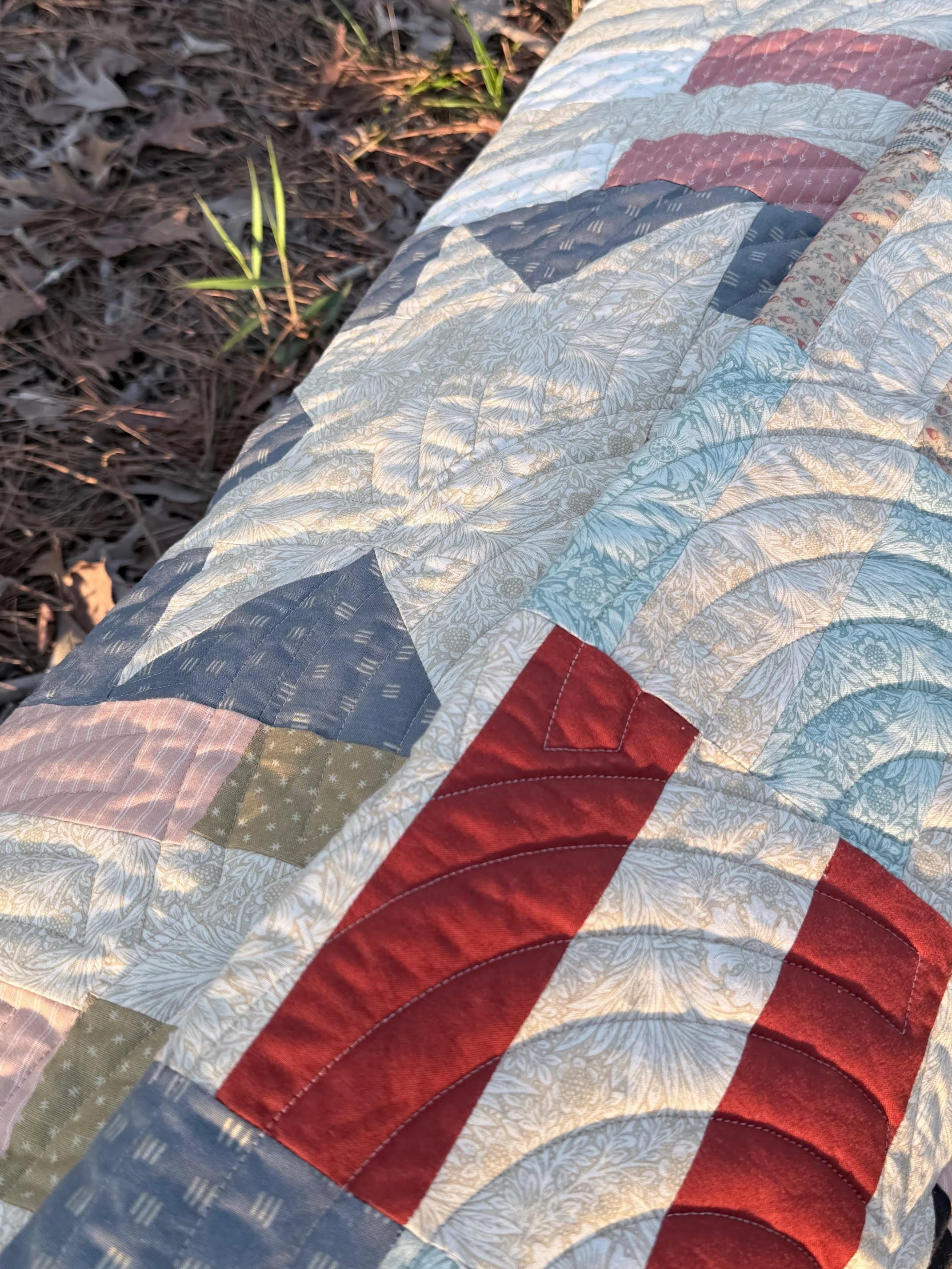 Close-up of a patchwork quilt with red, blue, cream, and patterned sections, lying on ground covered with dried leaves and grass.
