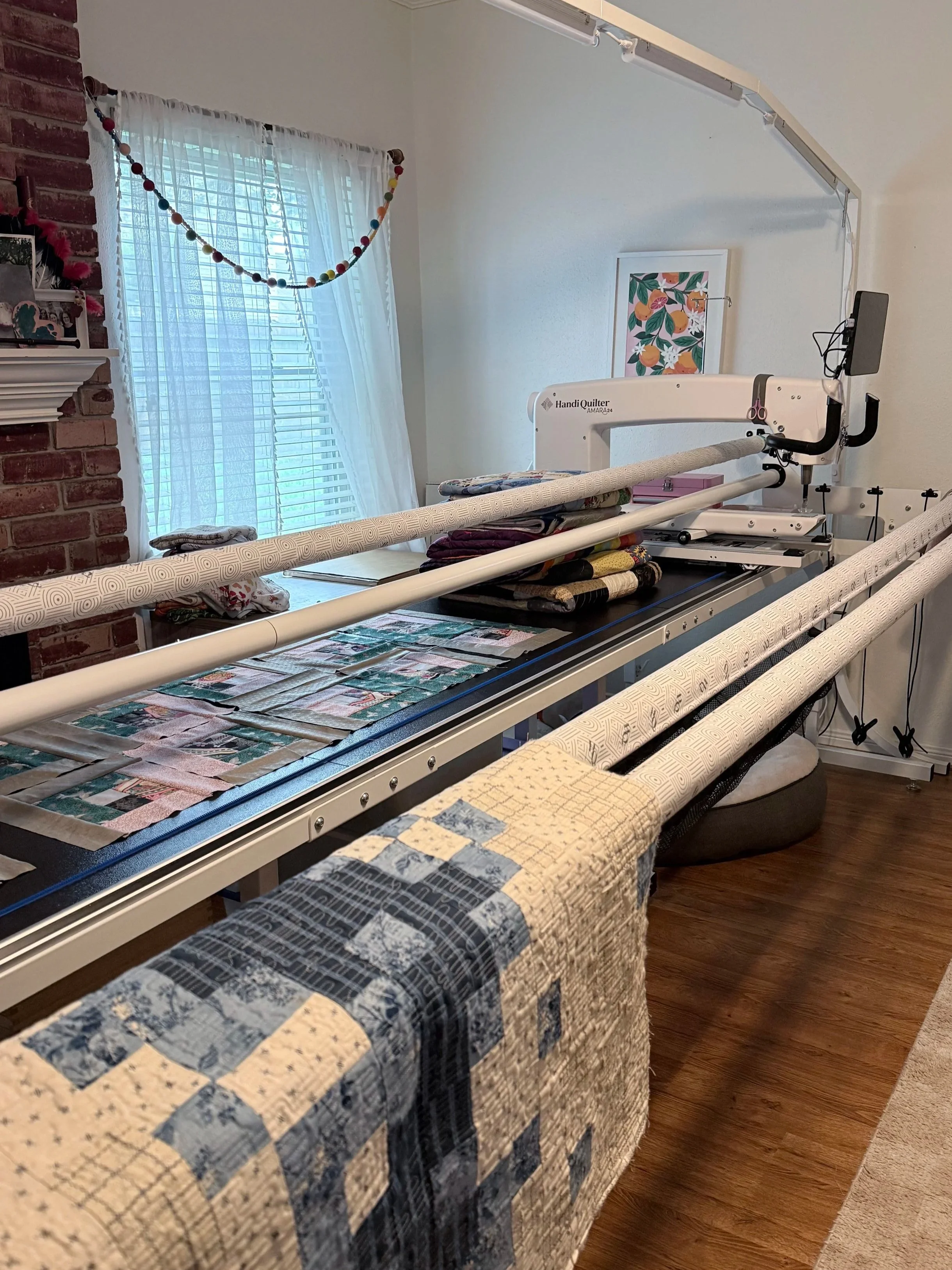 A quilting machine set up in a room with a window covered by sheer curtains, a string of colorful felt balls hanging, and various quilted fabric pieces on a table nearby.