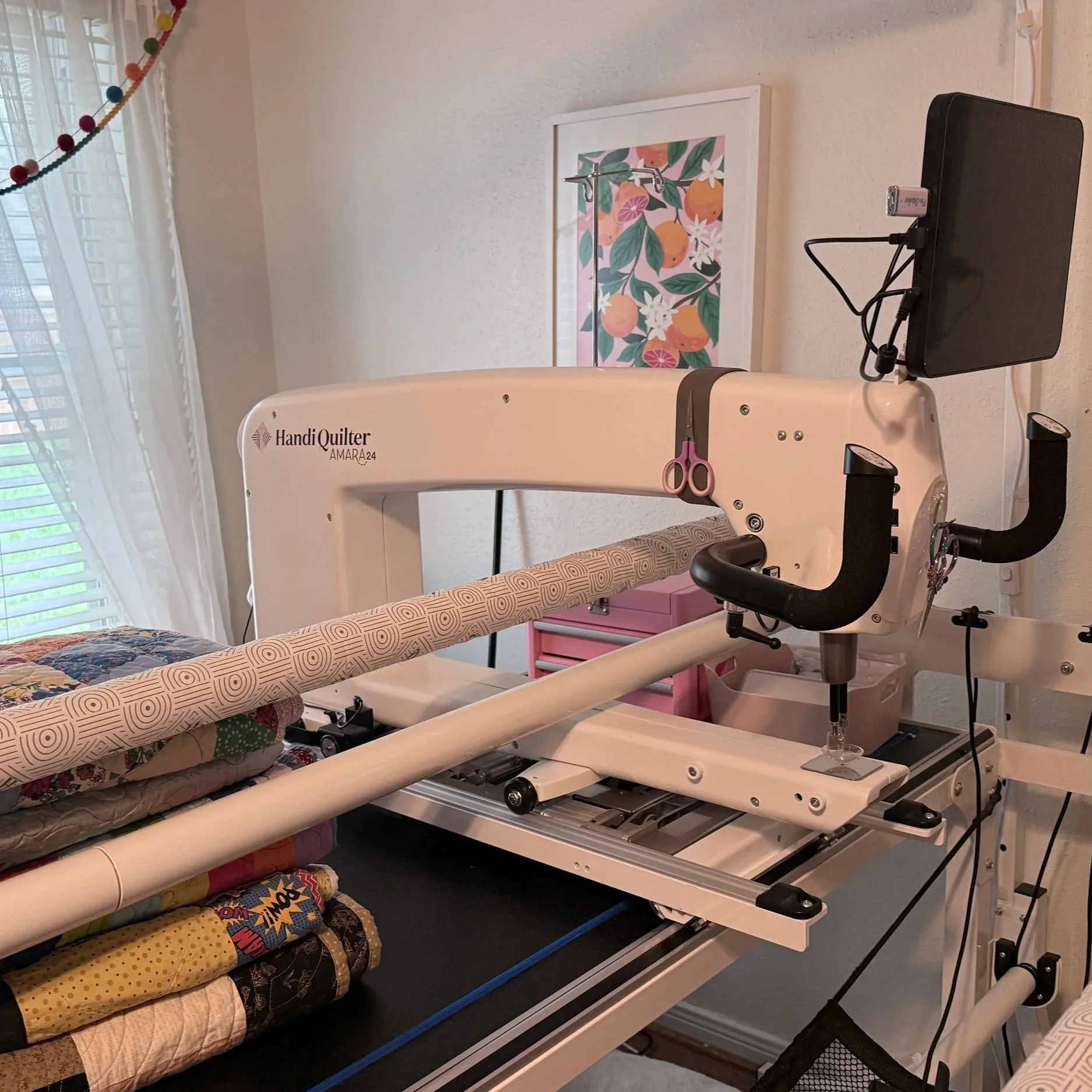 A machine, specifically a HandiQuilter AMARA quilting machine, positioned in a room with a bed and colorful blankets. Quilt fabrics are stacked beneath the machine, and the room has window blinds, a framed artwork of oranges, and a string of multi-colored decorations