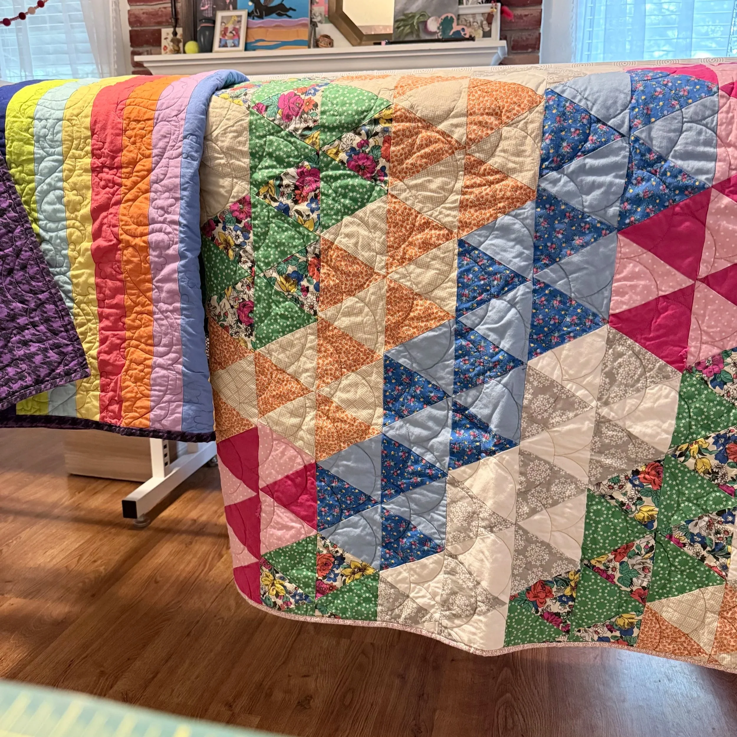 Colorful patchwork quilt with various floral and geometric patterns draped over a bed or furniture, with a brick wall and framed artwork in the background.