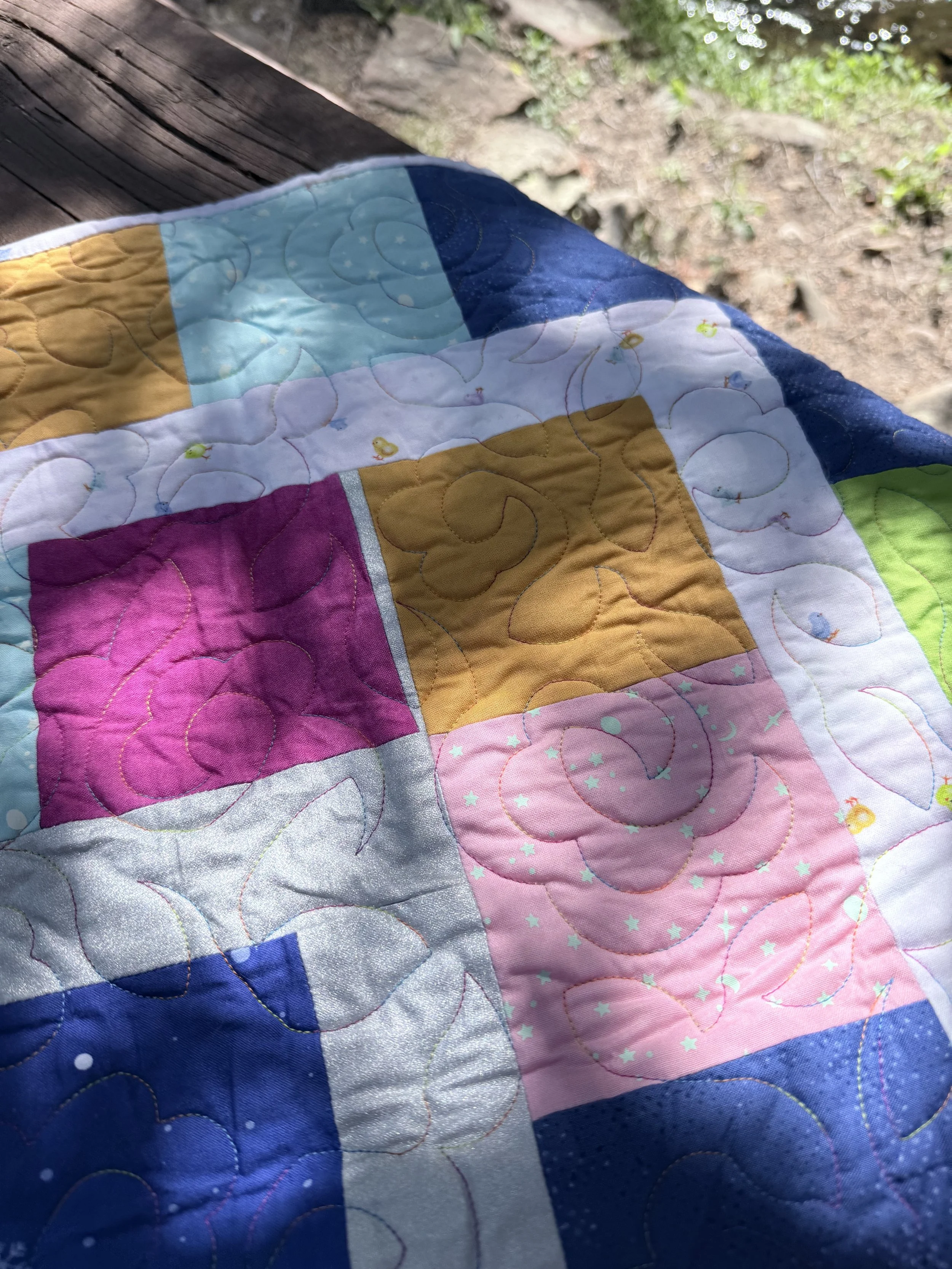 Colorful quilt with various patterns and colors, draped over a surface outdoors near dried leaves and grass.