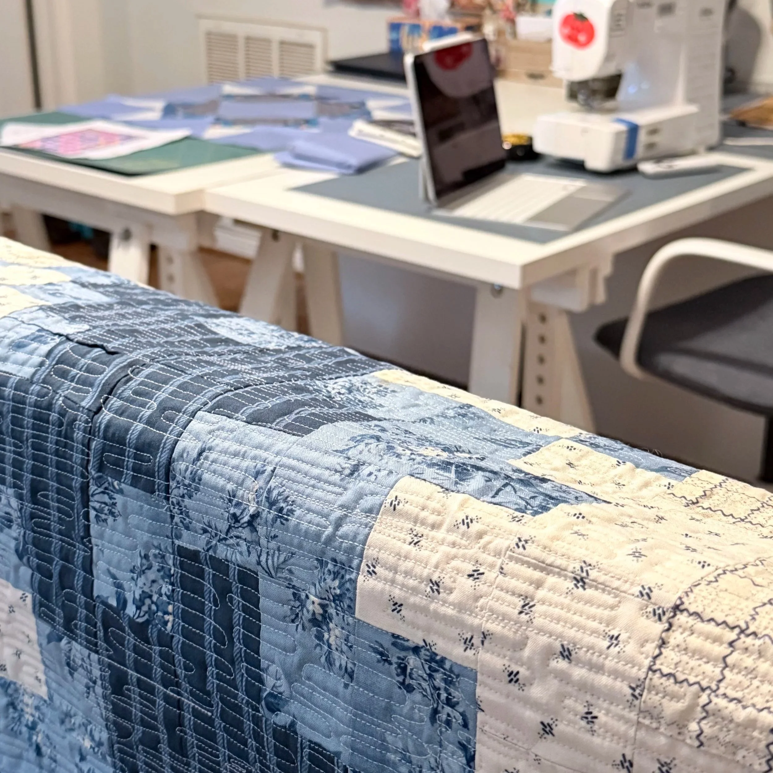 A desk with papers, a laptop, and office supplies in a quilt studio. 